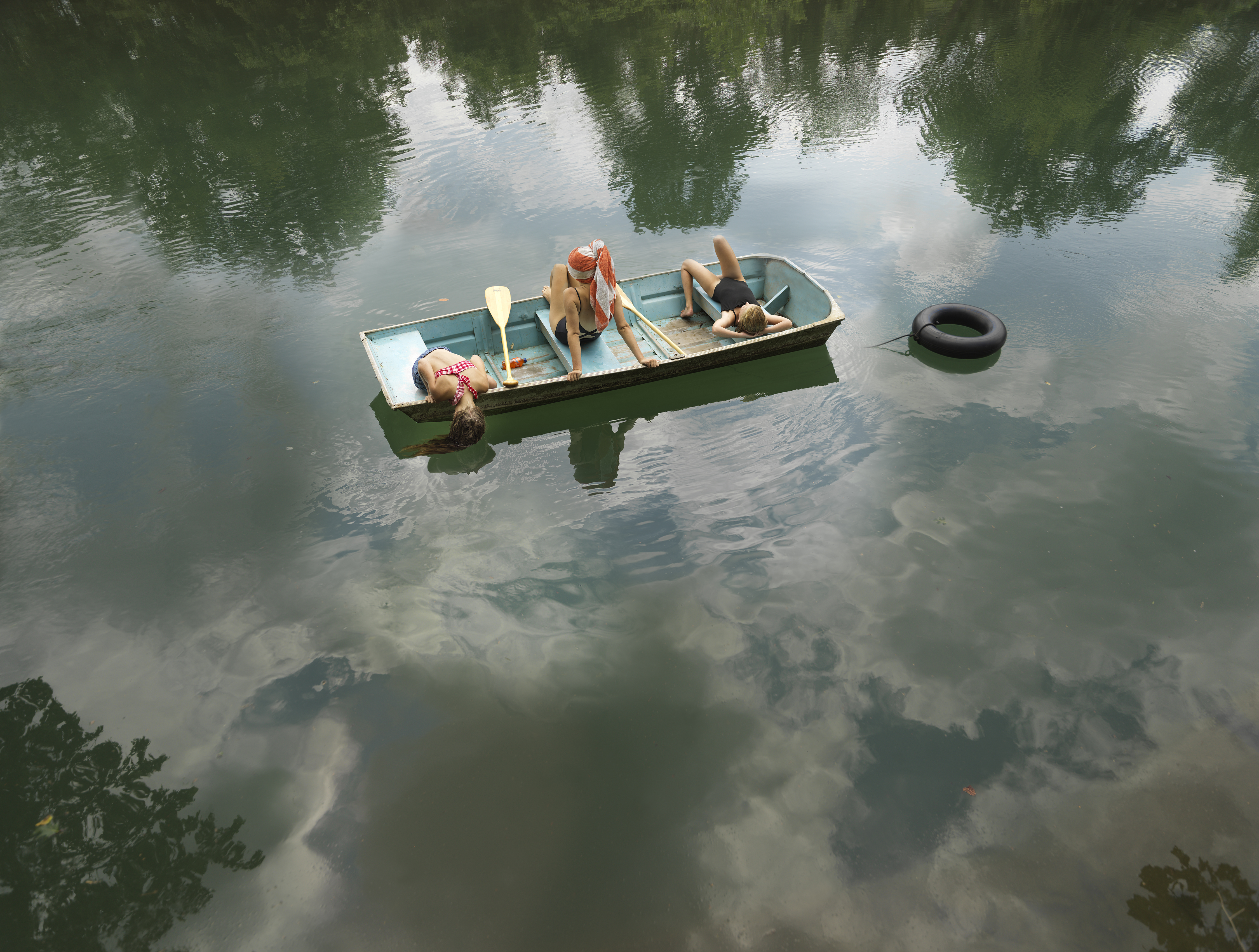 A small rowboat floats on calm, mirror-like water reflecting sky and surrounding trees. Three figures recline casually inside the boat among oars and supplies, while a black inner tube bobs in the water nearby. The serene composition captures a leisurely summer moment in muted greens and soft natural light.