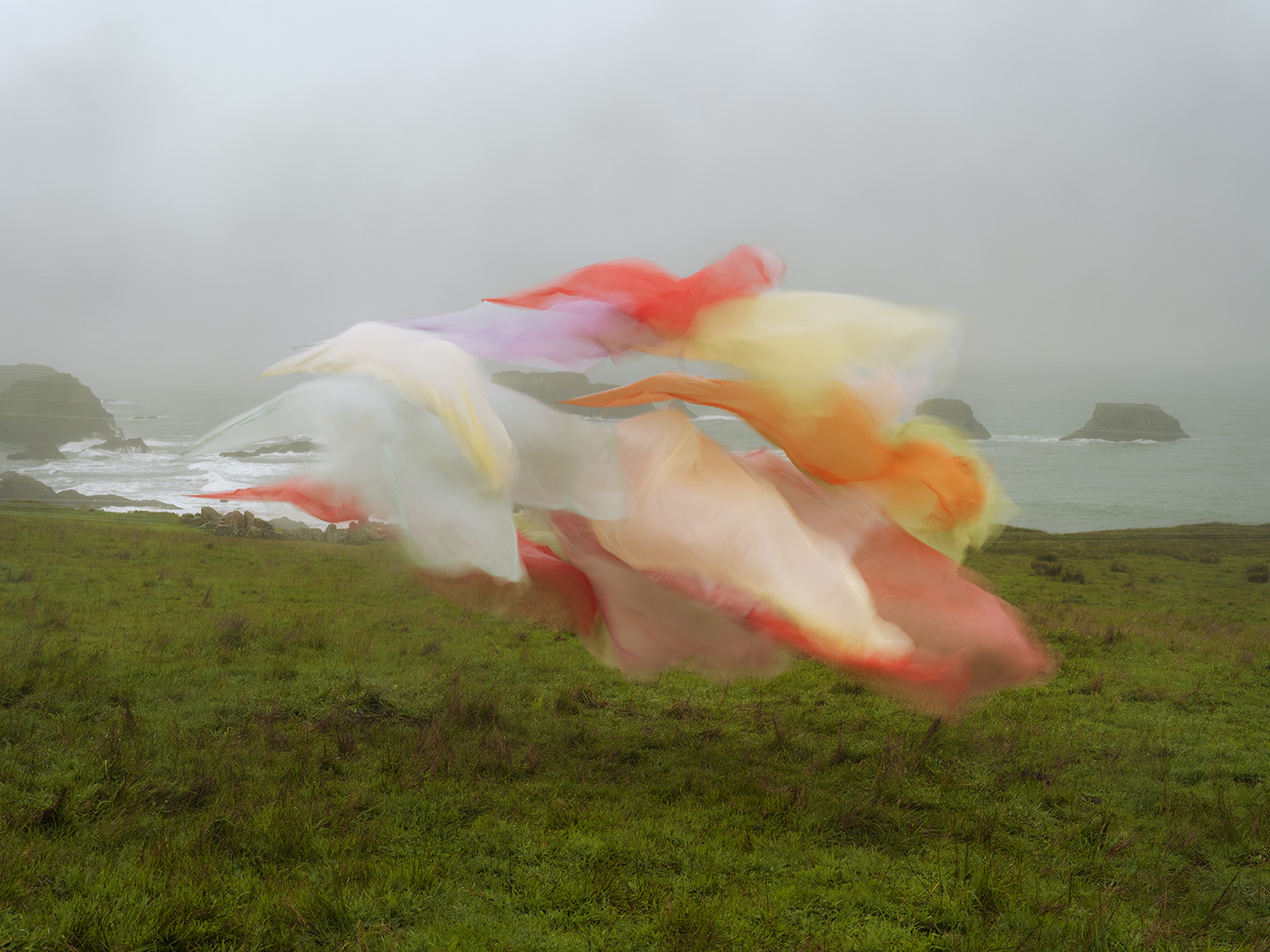 Colorful silk fabric in shades of red, orange, yellow, lavender, and white billows across a misty coastal meadow, with rocky outcroppings and turbulent sea visible in the fog-laden background.