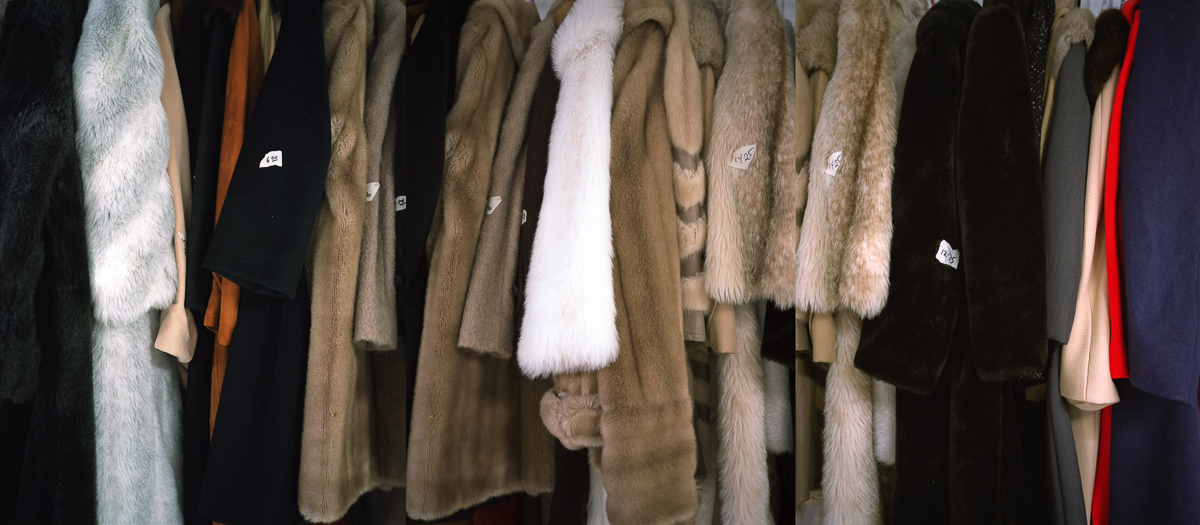 A row of fur coats and jackets hangs on display, featuring a range of neutral tones including cream, tan, brown, black, and white, with contrasting colored linings in orange and red visible at the edges. The garments vary in length and texture, creating a dense composition that emphasizes the luxurious quality and tactile nature of the furs.