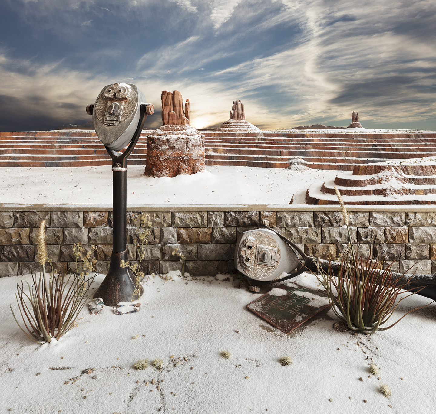 A snowy desert landscape with iconic red rock formations in the background beneath a dramatic cloudy sky. In the foreground, two vintage coin-operated telescopes stand on a stone wall overlooking the white-blanketed terrain, flanked by sparse desert vegetation and a weathered guidebook.