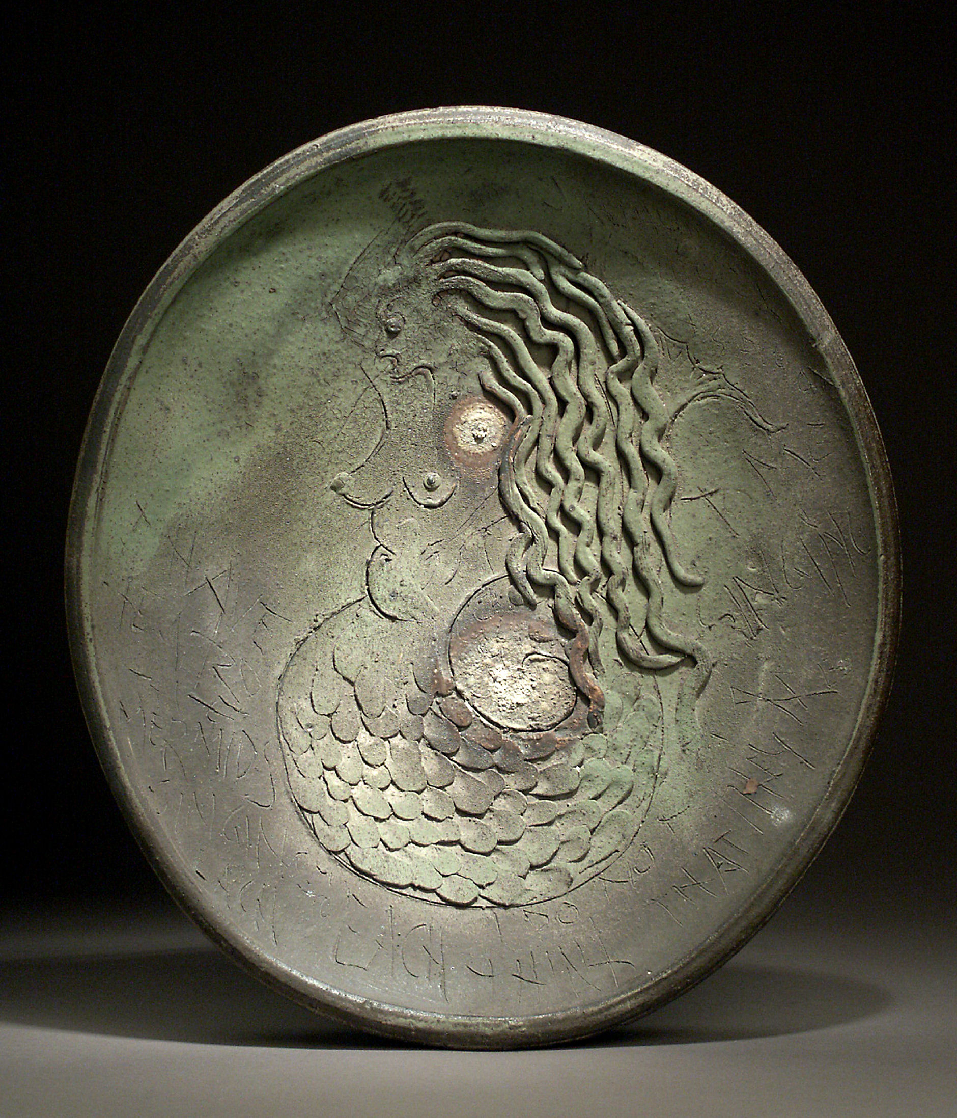 A relief sculpture on an oval platter depicts a mermaid in profile facing left, with long wavy hair and a scaled tail. The figure is rendered in dark stoneware with subtle metallic accents, creating a textured, three-dimensional composition against the recessed background.