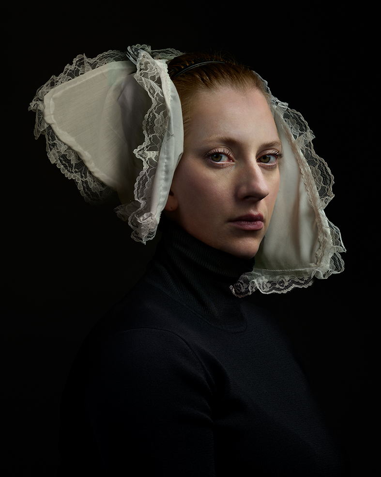 Portrait of a woman in profile against a black background, wearing a black turtleneck and an elaborate white linen bonnet with delicate lace trim and ties. The composition emphasizes the geometric structure of the historical headpiece and the subtle modeling of light across her face and neck.
