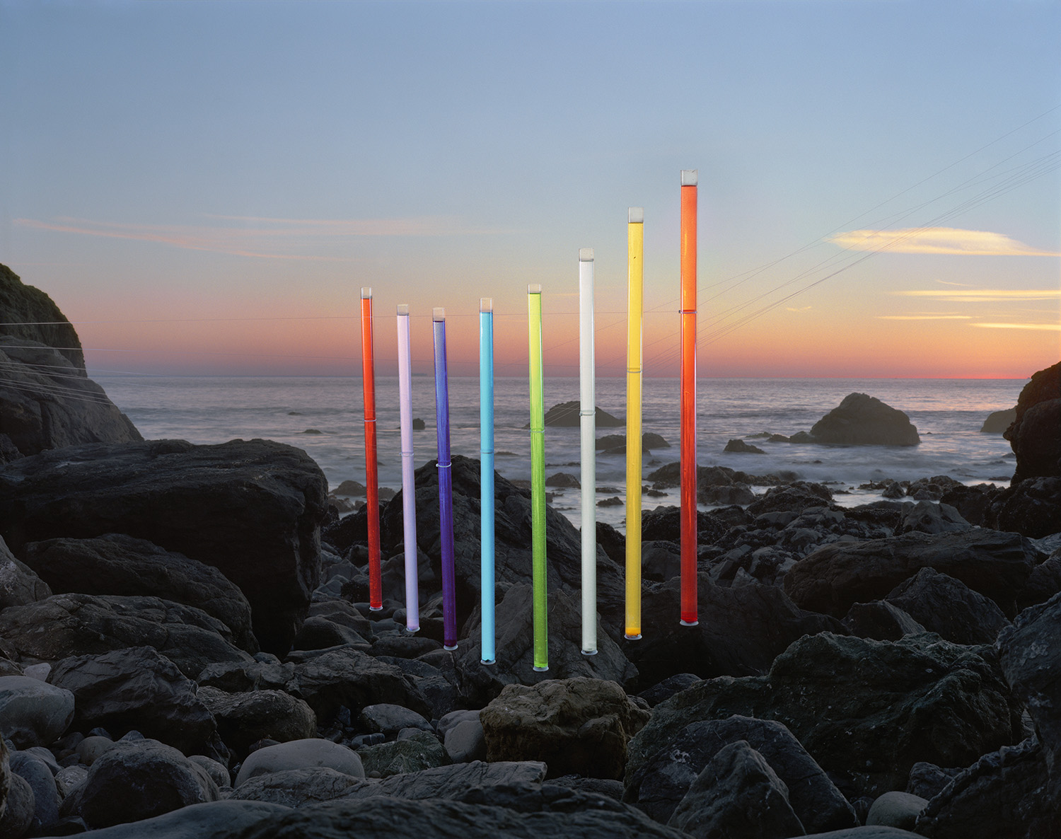 Nine illuminated neon tubes in graduated heights and colors—red, lavender, purple, cyan, lime, white, yellow, and orange—are installed among dark rocky outcroppings along a coastline at dusk, their bright glow contrasting against a soft pink and orange sunset sky over the ocean.