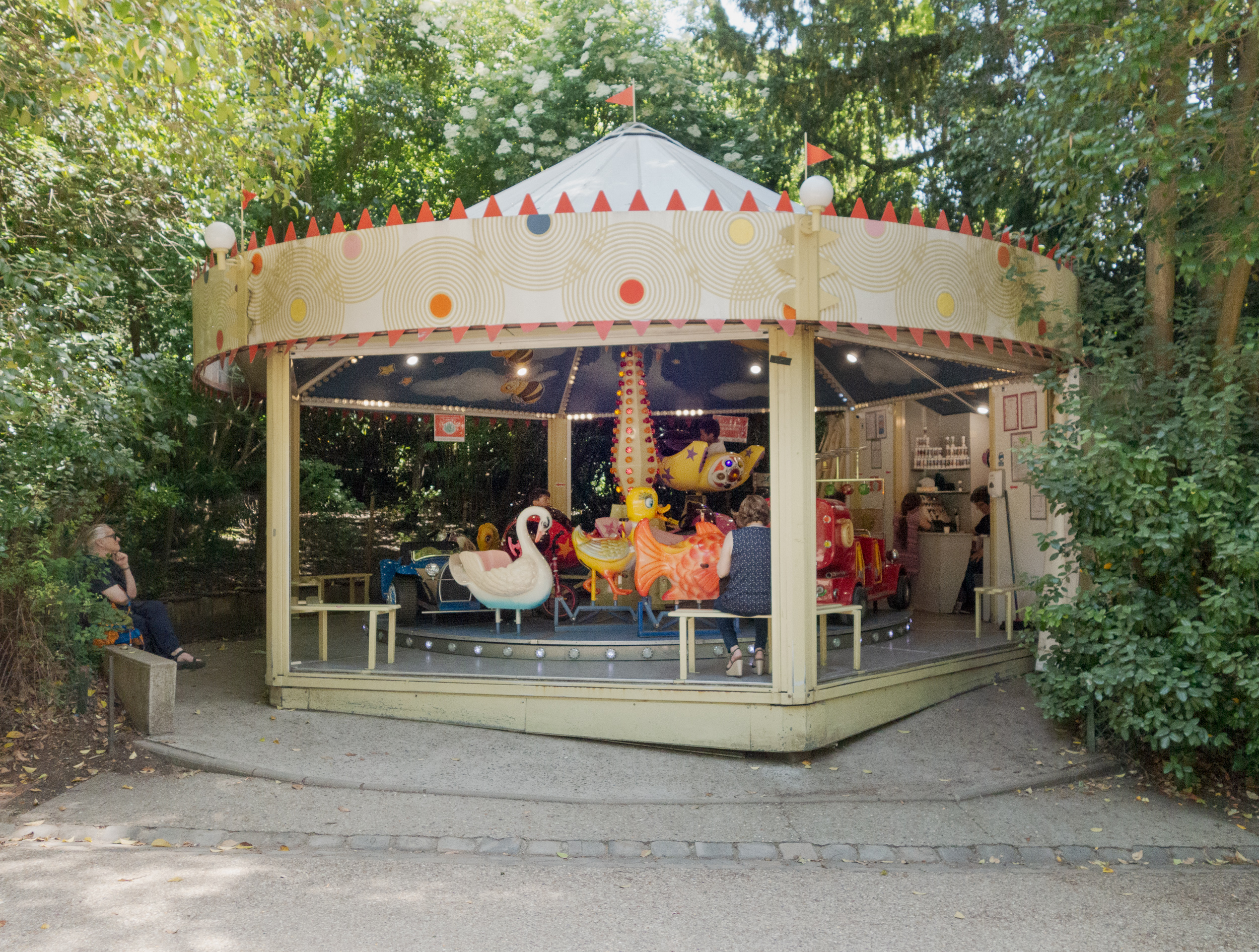 A whimsical carousel pavilion with a cream-colored cylindrical structure decorated with concentric circle motifs in muted tones and red triangular flags sits nestled among dense green foliage. The interior features colorful carousel animals including a swan and ornate horses, illuminated by warm lighting, with visitors visible through large windows.