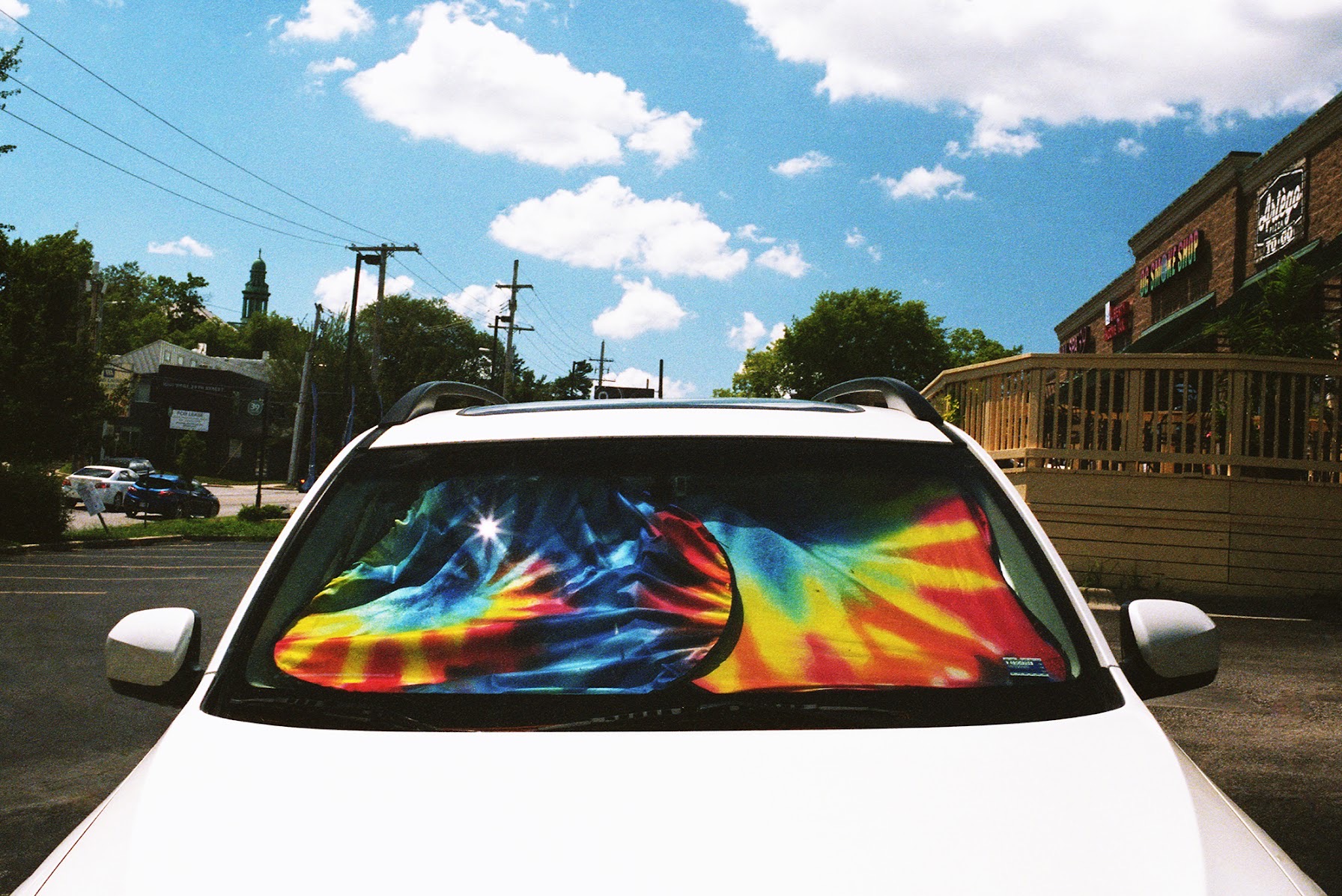 White vehicle parked on a city street with a vibrant, multicolored thermal-imaging style sunshade displayed across its windshield, featuring intense reds, yellows, blues, and greens in an abstract pattern. The surrounding urban landscape with brick buildings, utility poles, and cloudy sky is visible in the background.