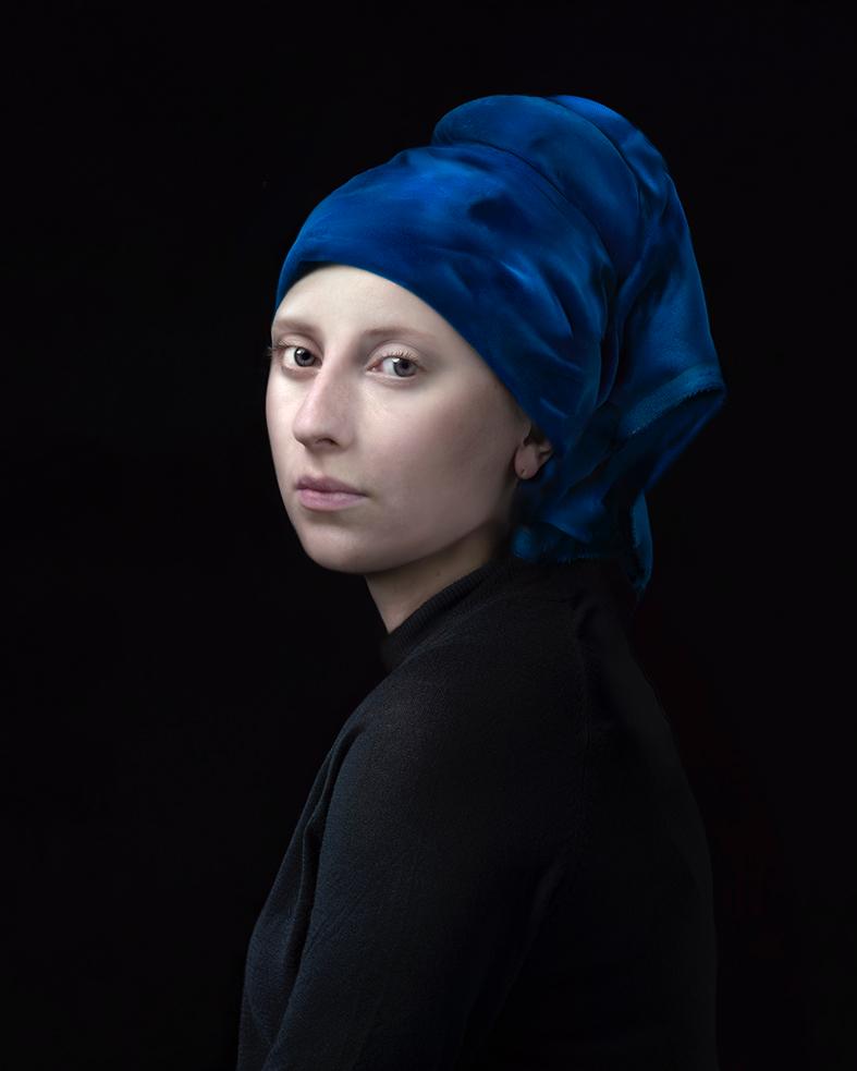 A figure with pale skin wearing a voluminous blue turban and black garment turns to gaze over their shoulder against a dark background, rendered in a contemporary portrait style with striking color contrast and subtle modeling of form.