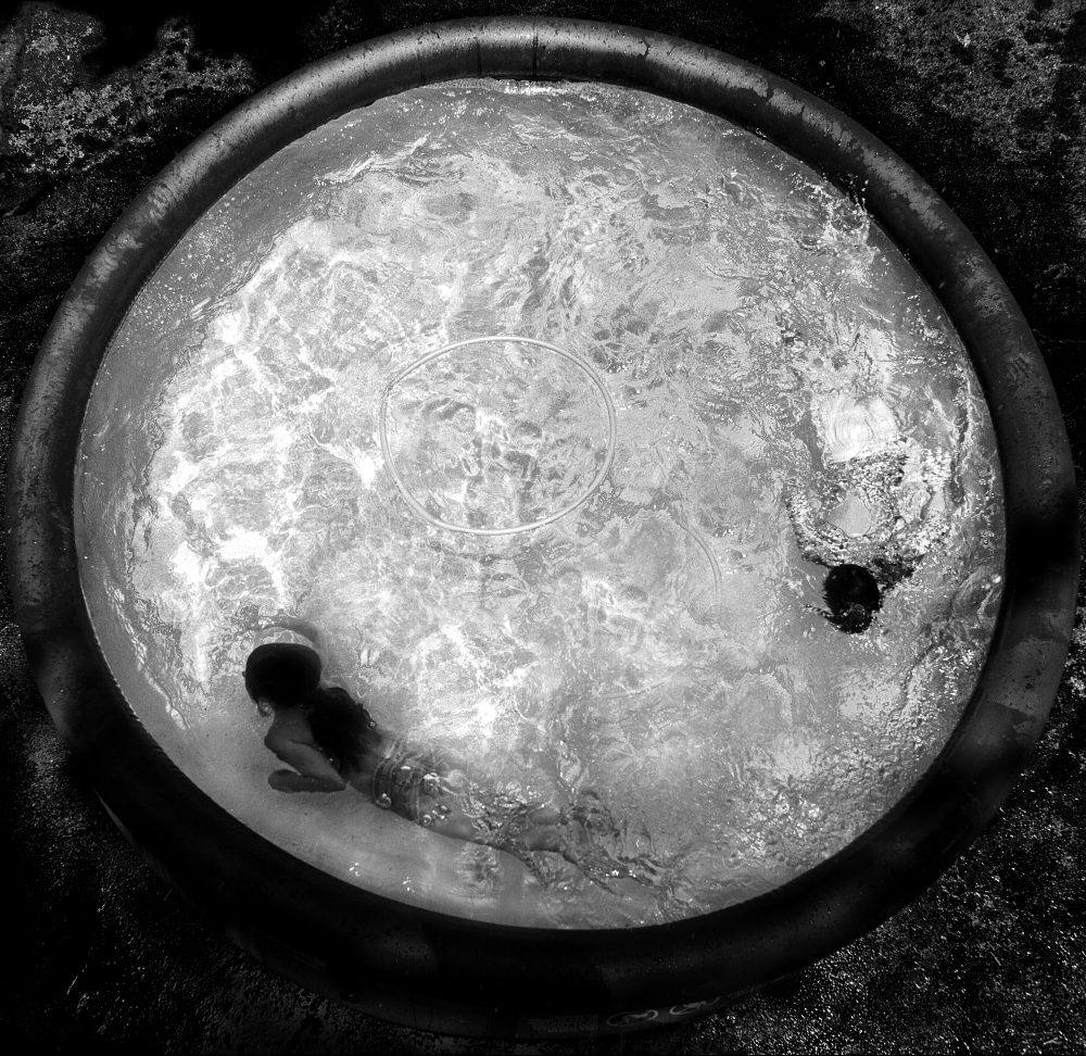 Black and white overhead view of a child floating in a shallow circular pool with textured water surface, surrounded by dark ground. Two dark voids appear at the bottom and right side of the pool's interior.