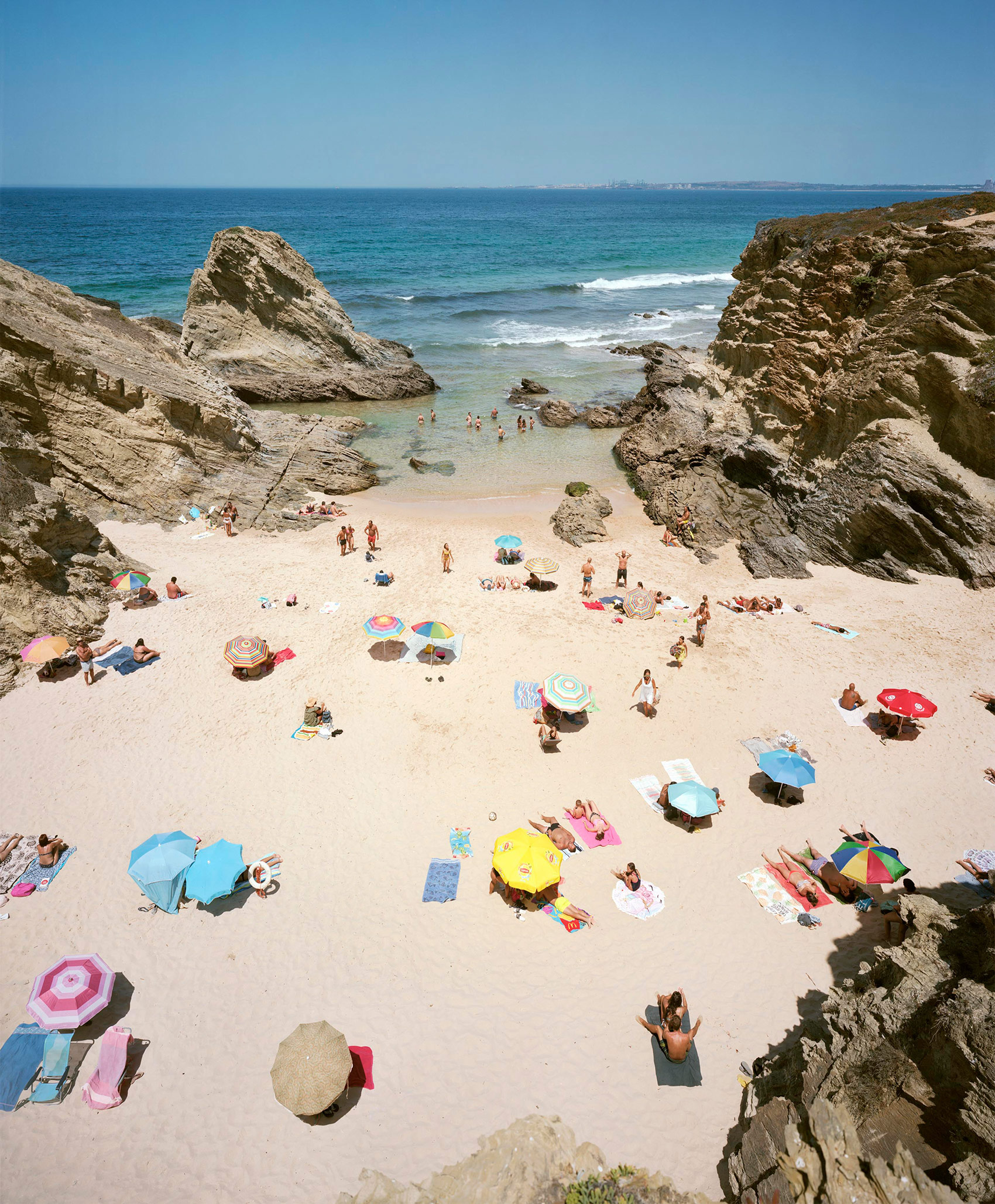 Aerial view of a crowded cove beach framed by dramatic limestone cliffs, with colorful umbrellas and beachgoers scattered across pale sand, turquoise water, and rocky outcrops under clear blue sky.