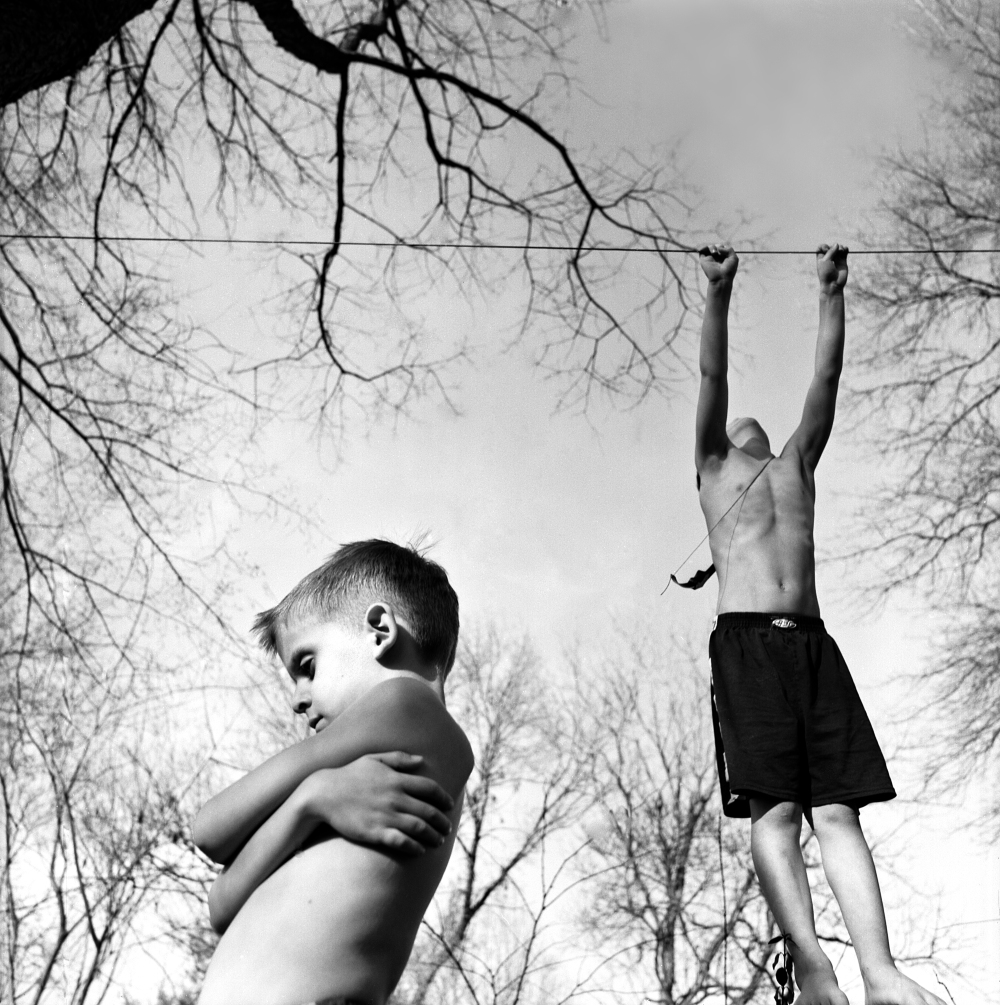 Black and white photograph of two shirtless boys outdoors beneath bare tree branches. One boy in the foreground looks downward with his arms crossed over his chest, while the second boy hangs from a horizontal wire or rope stretched across the composition, his arms extended upward. The overcast sky and leafless trees create a stark, somber atmosphere.