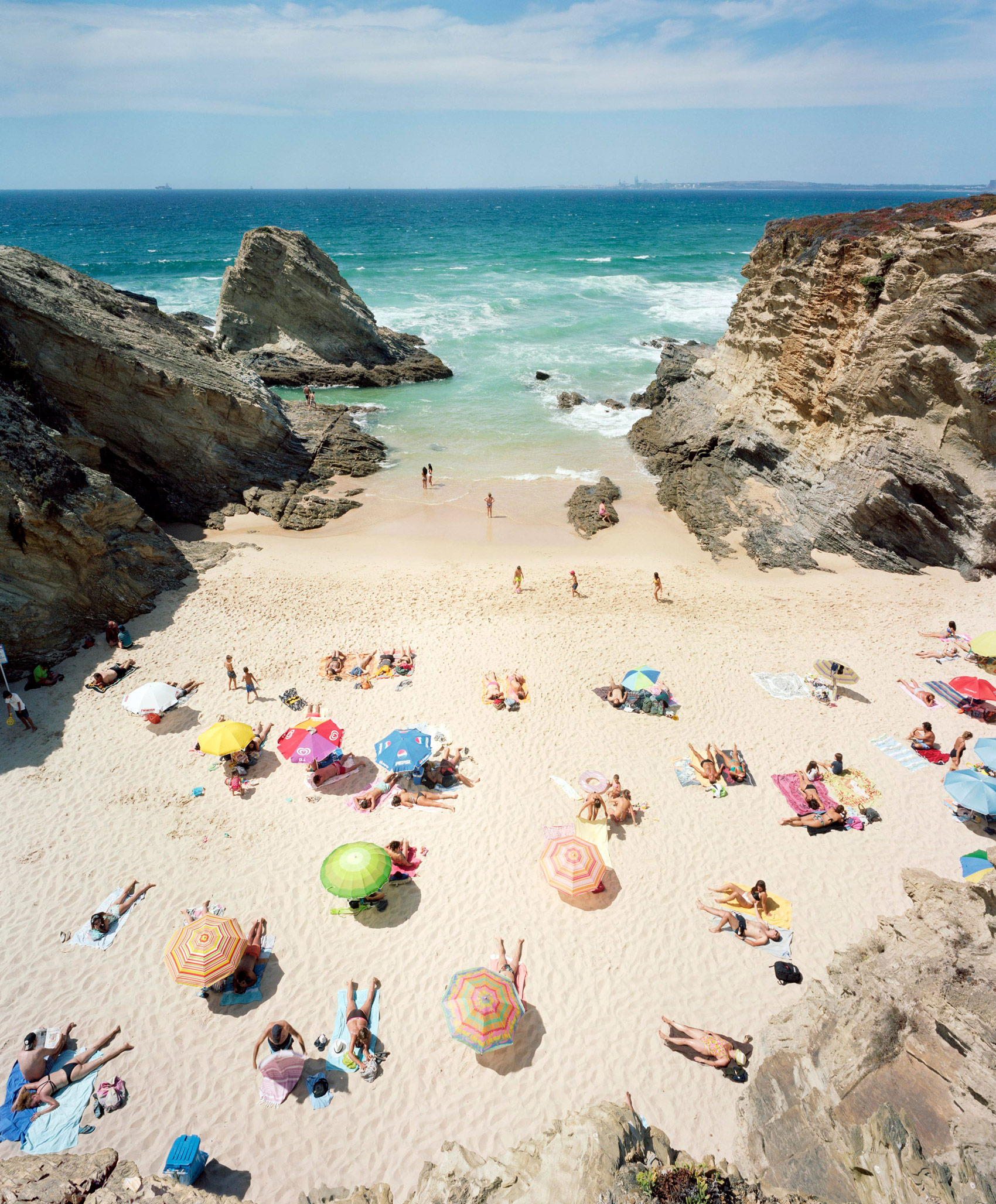 Aerial view of a crowded beach cove framed by dramatic rocky cliffs, with numerous sunbathers scattered across pale sand beneath colorful umbrellas in yellow, pink, blue, and green, while turquoise waves and white foam lap at the shoreline.