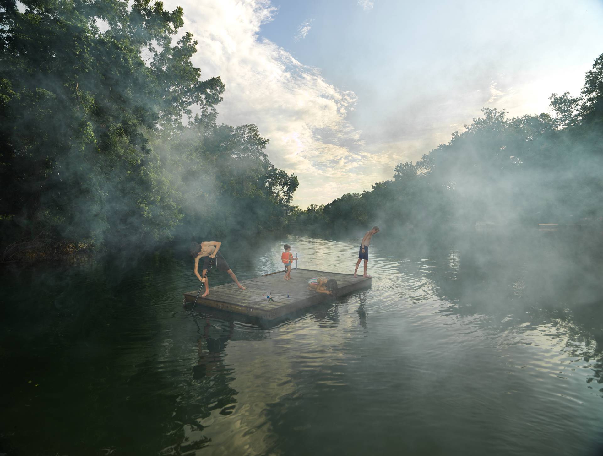 Four figures on a wooden raft float in a misty river surrounded by dense green forest, with morning mist rising from the water's surface and dramatic clouds visible through the canopy above.