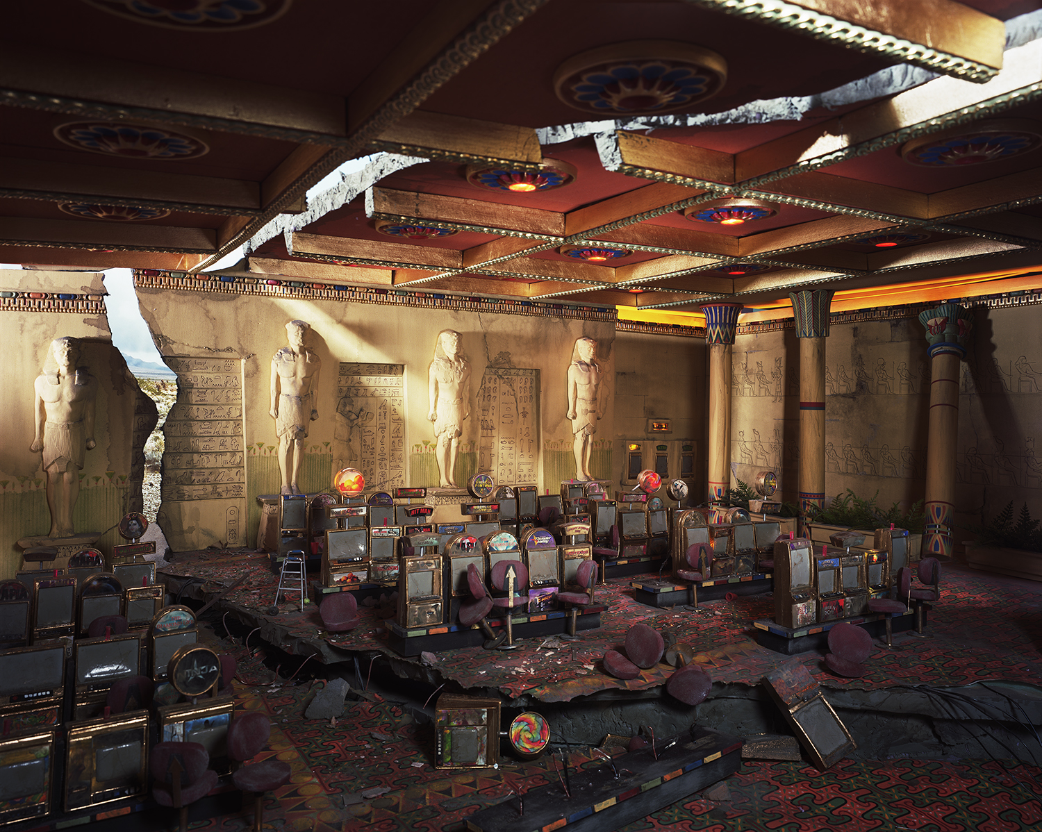 An abandoned casino interior with Egyptian-themed décor featuring colossal statues, hieroglyphic-covered walls, and ornate coffered ceilings. Rows of slot machines in burgundy and gold tones are scattered across a patterned carpet, with debris and overturned chairs throughout the deteriorating space.