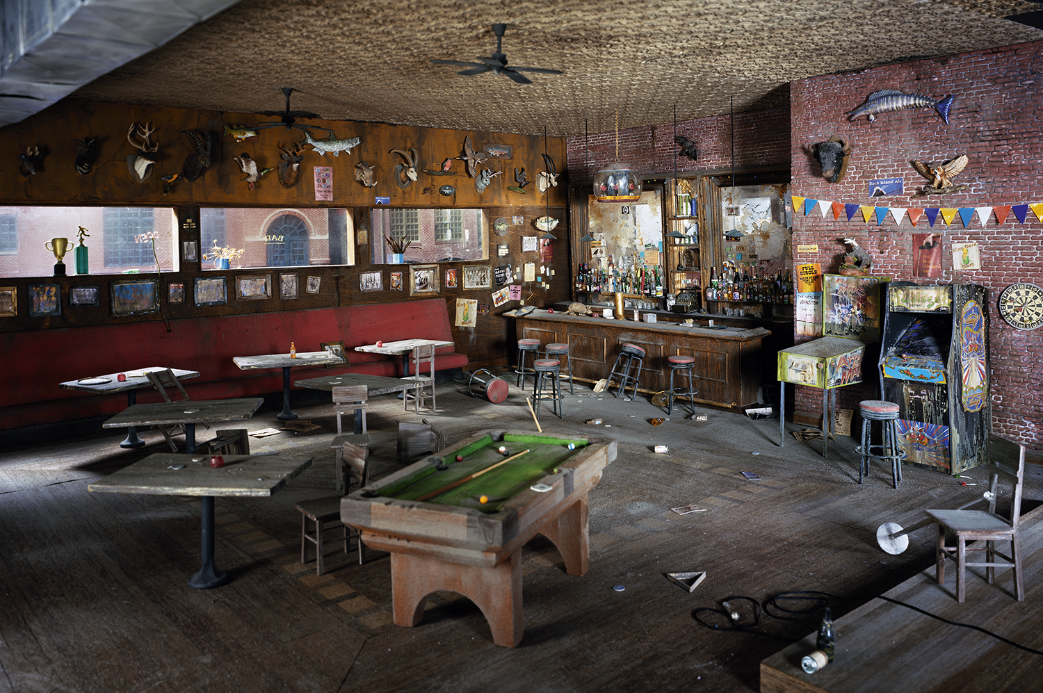 An abandoned interior recreation of a dimly lit bar and recreation room, featuring a pool table in the foreground, a wooden bar counter with shelves of bottles, red vinyl booth seating, taxidermied animal heads mounted on wood-paneled walls, vintage arcade games, and miscellaneous memorabilia scattered throughout. Exposed brick, concrete floors, and ceiling fans create an atmosphere of urban decay and nostalgia.