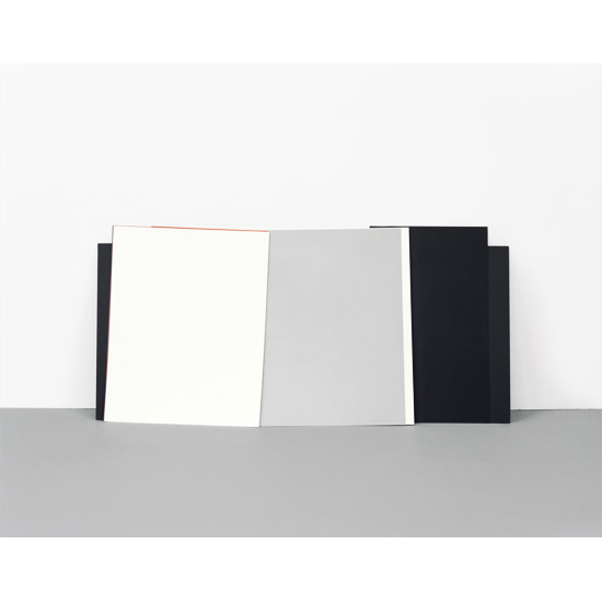 Four overlapping rectangular panels in varying colors—black, cream, light gray, and dark charcoal—lean against a gray surface against a white wall, creating geometric shadows and a minimalist compositional study.