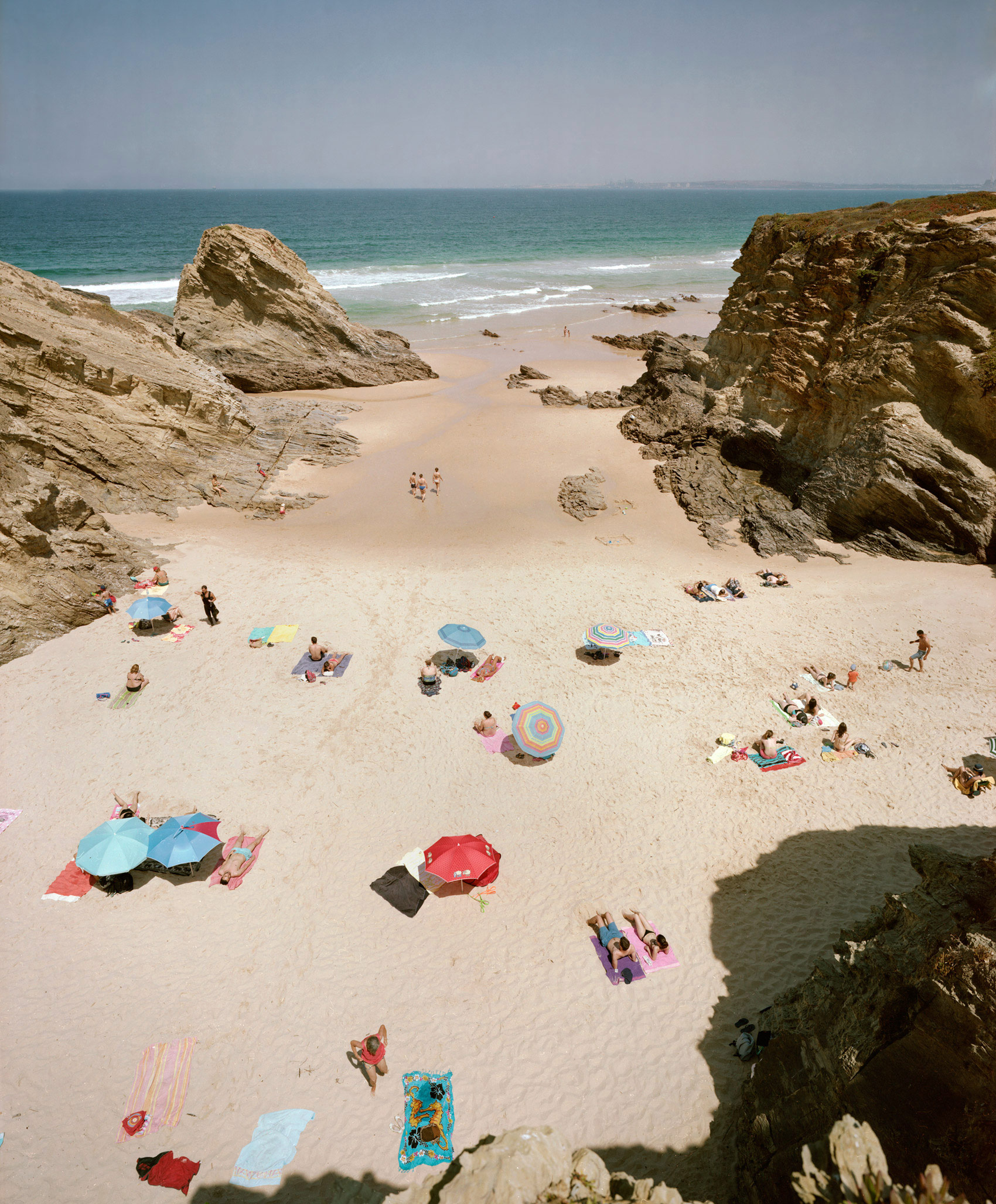 Aerial view of a secluded beach cove framed by dramatic rocky outcrops, with beachgoers scattered across pale sand beneath colorful umbrellas and towels, turquoise waves visible in the distance.