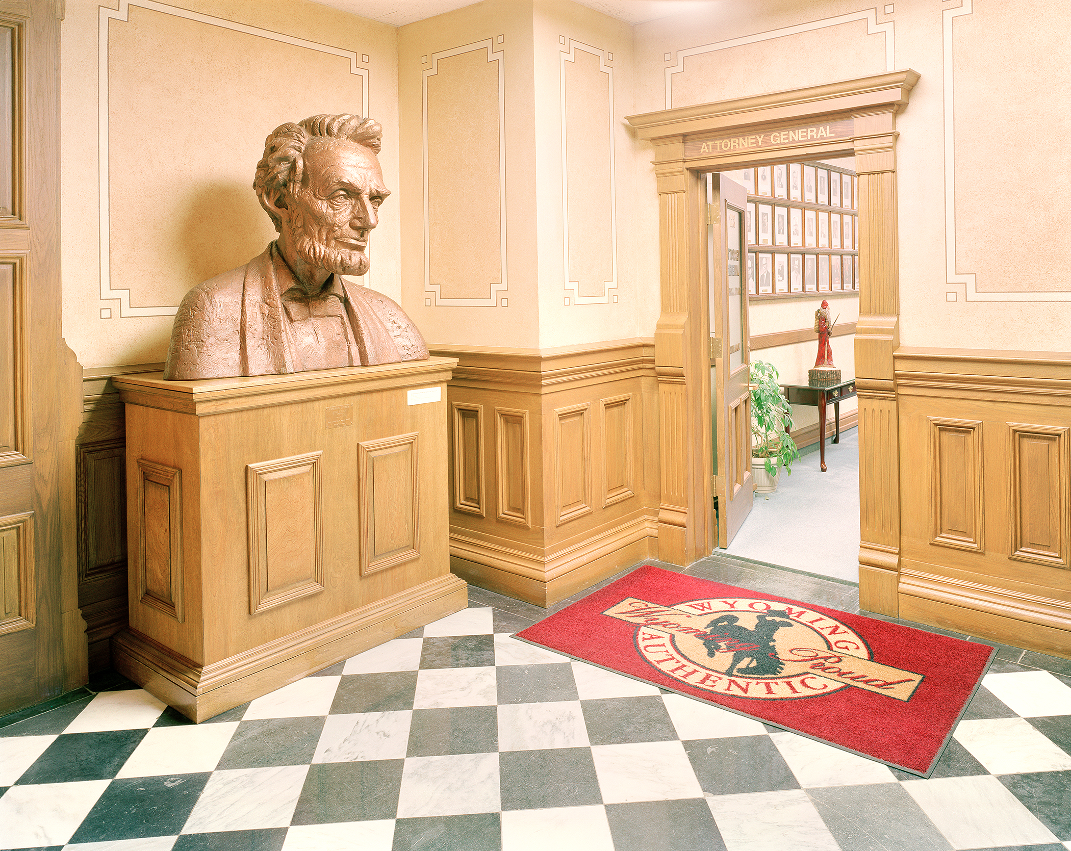A bronze bust of a bearded historical figure sits on a wooden pedestal in a classical interior hallway with cream-colored walls trimmed in geometric patterns. The space features warm wood paneling, a black-and-white diamond-patterned floor, and an "Attorney General" doorway visible in the background, with a red mat bearing an official seal in the foreground.