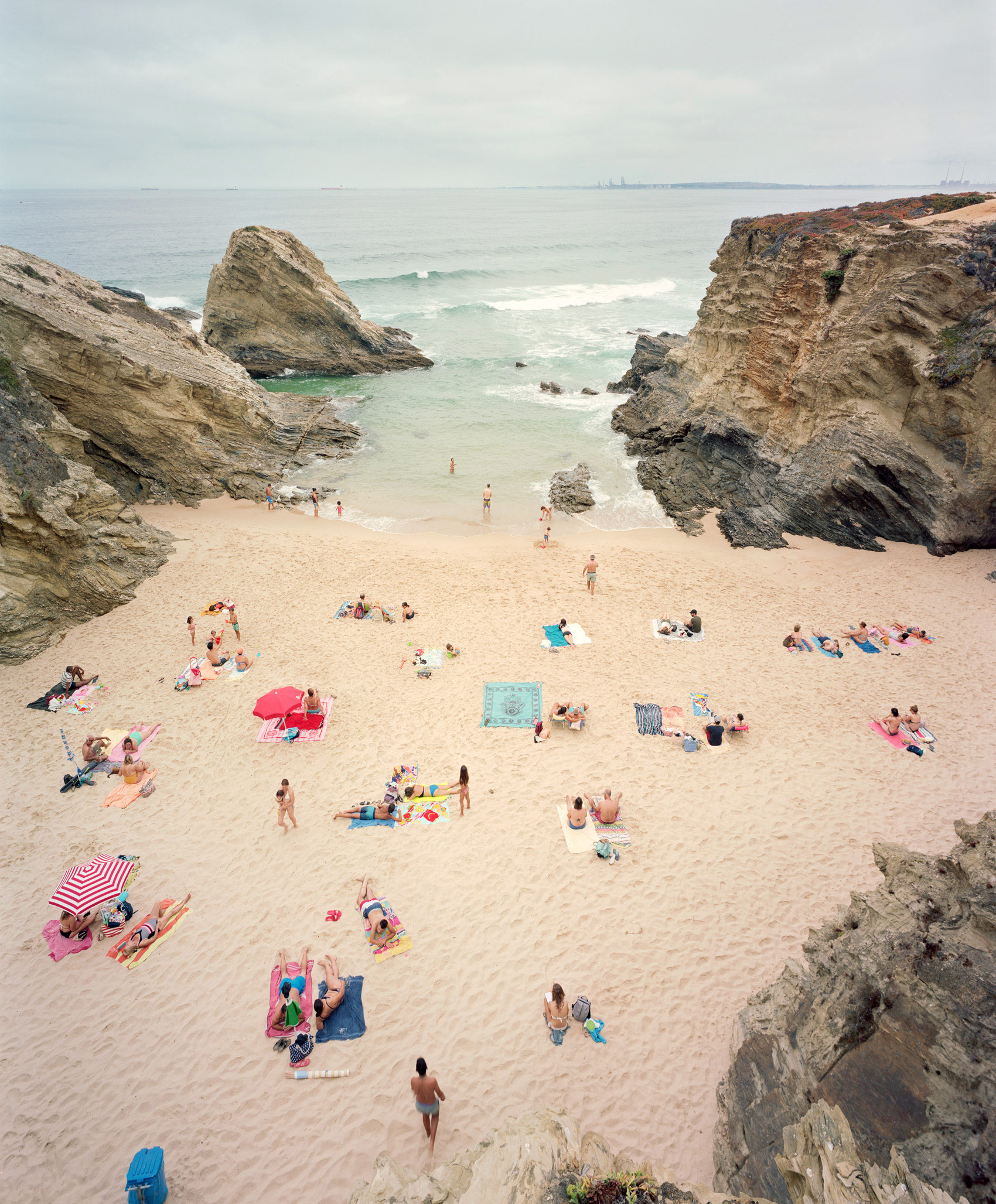 Aerial view of a sandy beach cove enclosed by dramatic rocky cliffs, with scattered beachgoers, colorful umbrellas, and towels dotting the sand. The turquoise and white waters beyond are studded with rocks, under an overcast sky.