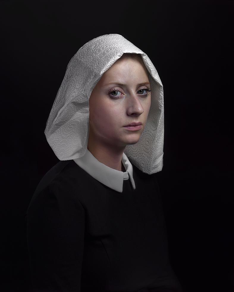 Portrait of a young woman in three-quarter view against a black background, wearing a black garment with white collar and an elaborate white linen headdress with voluminous folds. Her gaze is directed downward and to the side with a contemplative expression.