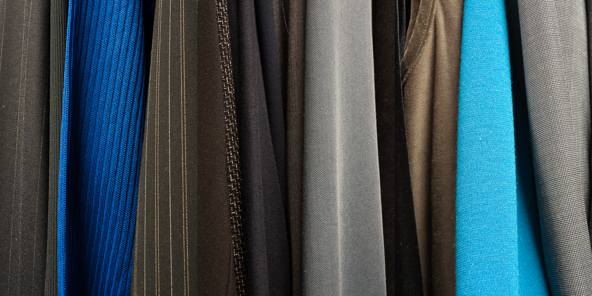 Vertical stripes of fabric in varying textures and colors—charcoal, deep blue, black, gray, tan, and bright cyan—hang closely together creating an abstract composition that emphasizes material contrast and subtle color gradations.