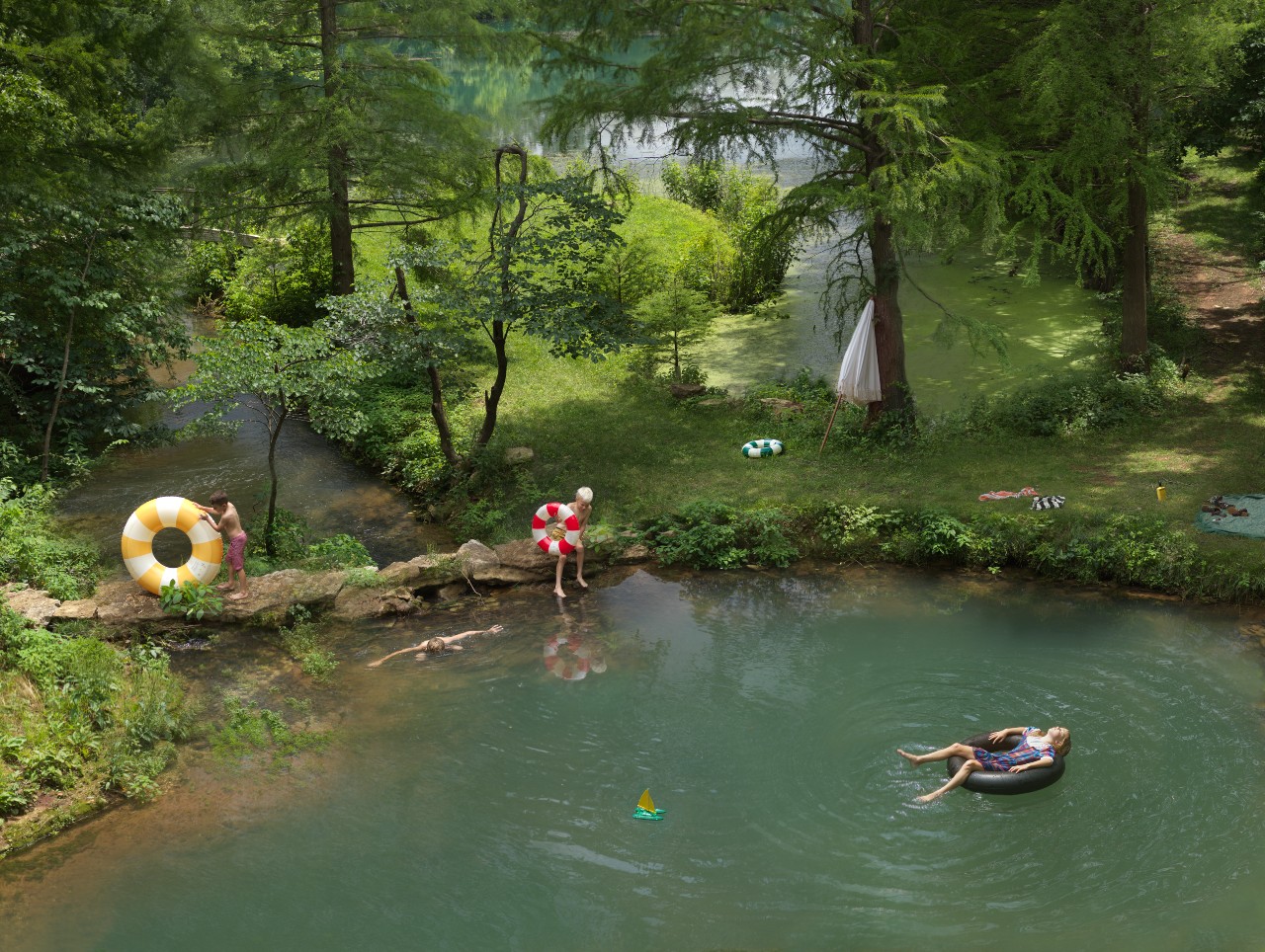 Aerial view of a lush forest landscape with a clear green river running through it. Several people engage in summer leisure activities: a child in a red and white inner tube stands on the riverbank beside a yellow and white float ring, while a person floats on an inflatable raft in the water. Additional figures rest on the grassy banks among towering trees, and a beige umbrella provides shade in the distance.