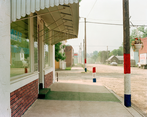 A small-town storefront with brick base and white upper walls sits beneath a green awning, its window reflecting surrounding buildings. The perspective extends down an empty main street lined with utility poles and painted bollards in red, white, and blue, with rural structures visible in the hazy distance.