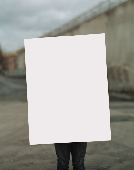 A person stands outdoors holding a large blank white rectangular sheet in front of their body, obscuring their face and torso. Behind them, an industrial waterfront landscape with concrete structures and overcast sky is softly blurred.