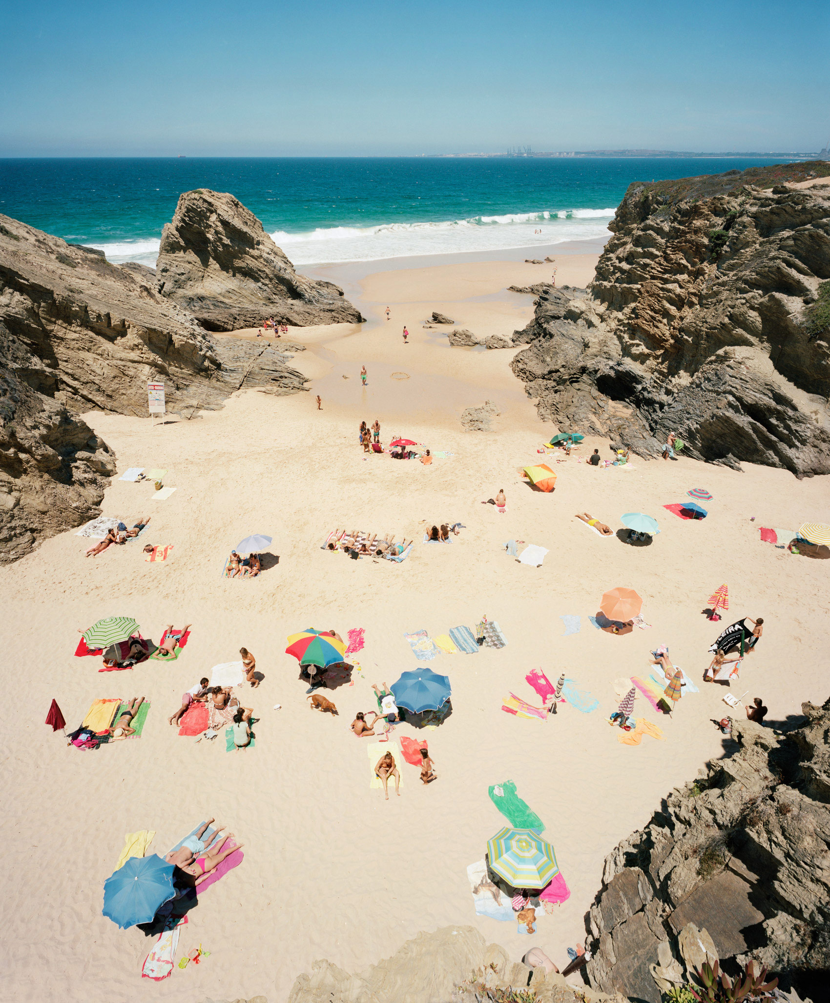 Aerial view of a secluded beach cove framed by dramatic rocky outcroppings, with numerous sunbathers scattered across the sand beneath a vibrant array of colorful beach umbrellas and towels in shades of pink, blue, yellow, and green. Turquoise waves break along the shoreline while the horizon extends to a distant coastline under clear blue sky.