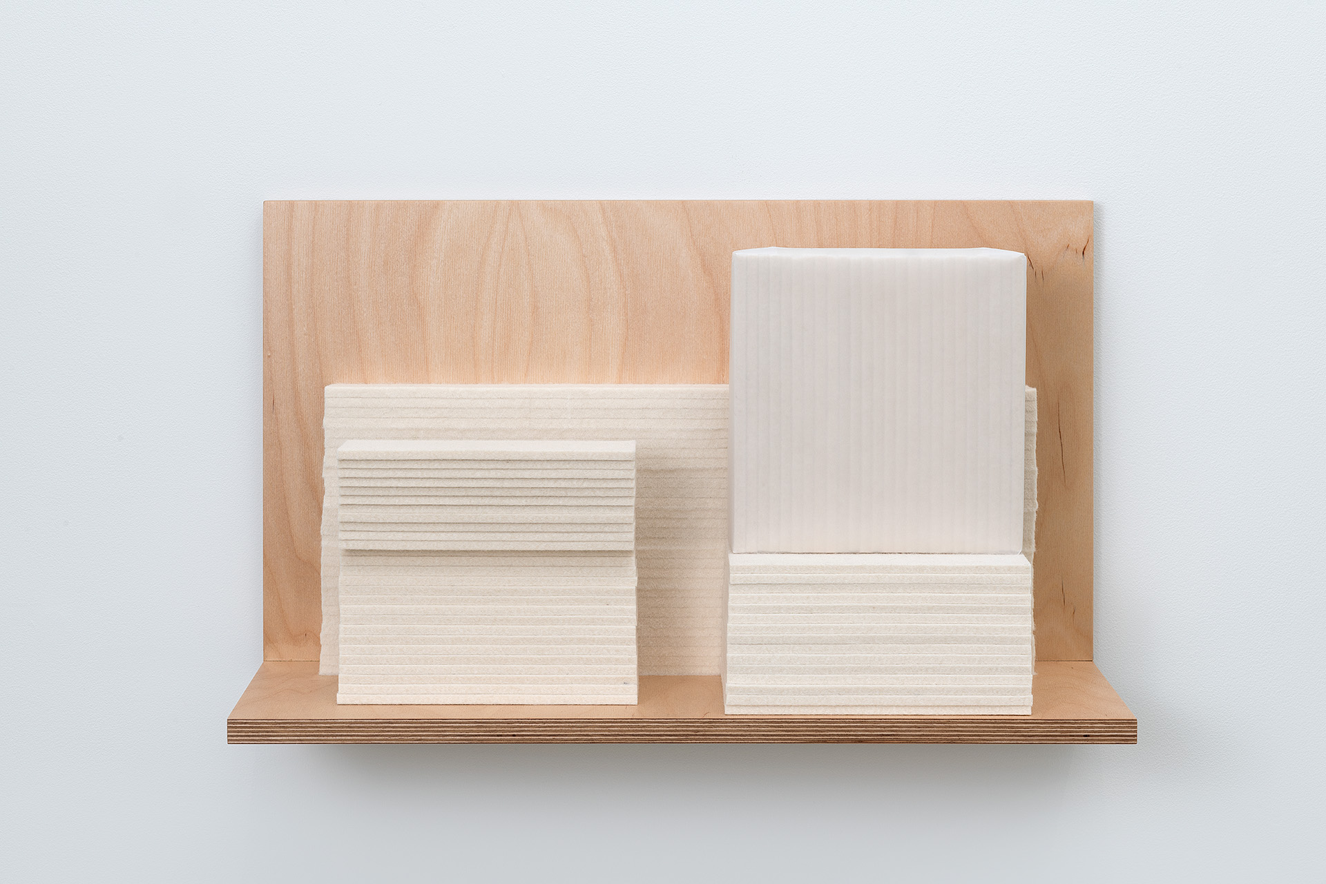 A wooden shelf mounted on a light gray wall holds stacked rectangular forms made of cream-colored wool felt arranged in two groups. The felt pieces are layered horizontally, creating subtle linear textures across their surfaces, while the natural plywood shelf and backing provide warm contrast to the soft, matte finish of the materials.