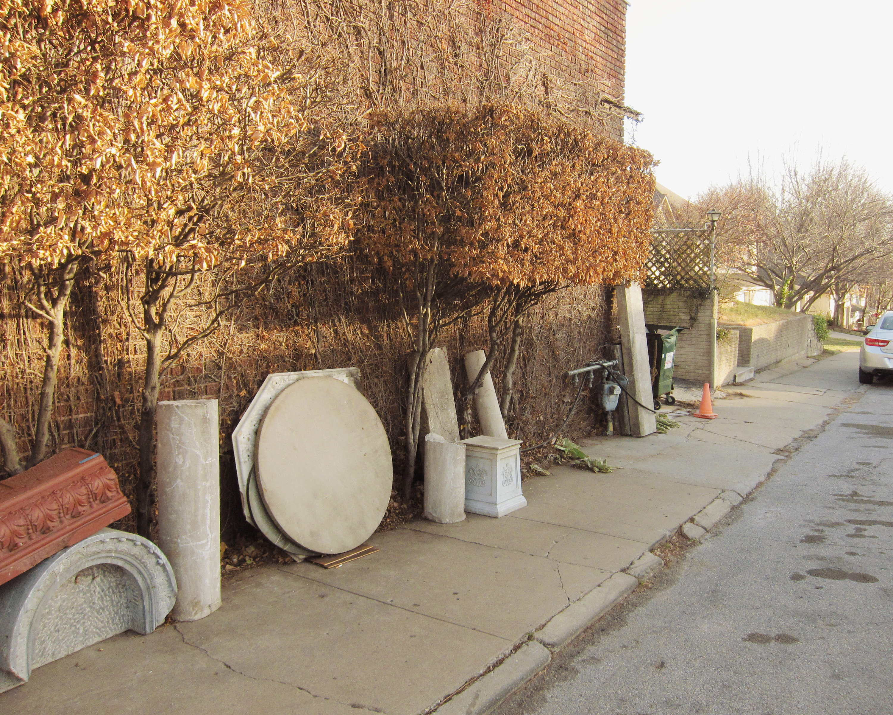 Alleyway lined with ivy-covered brick walls and dormant vines, featuring discarded architectural salvage including large concrete pipes, a round white basin, decorative molding, and other building fragments arranged along a concrete sidewalk.