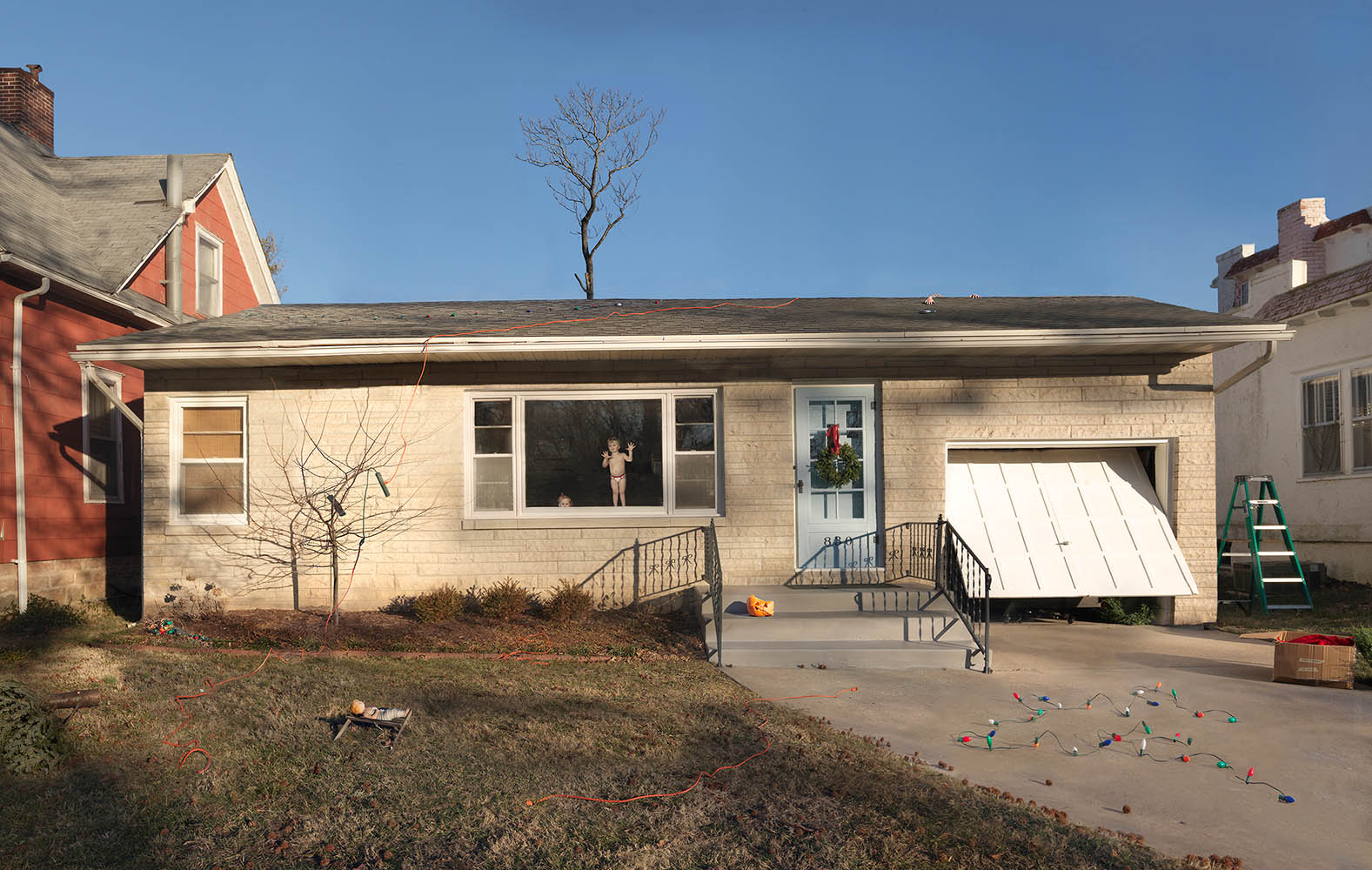 A residential house with cream-colored brick exterior is photographed from the front, featuring a central door with a wreath, large windows, and a white awning over the garage. A figure stands visible through the central window, while toys and decorations scatter across the concrete driveway and sparse lawn, adjacent to neighboring houses under a clear blue sky.