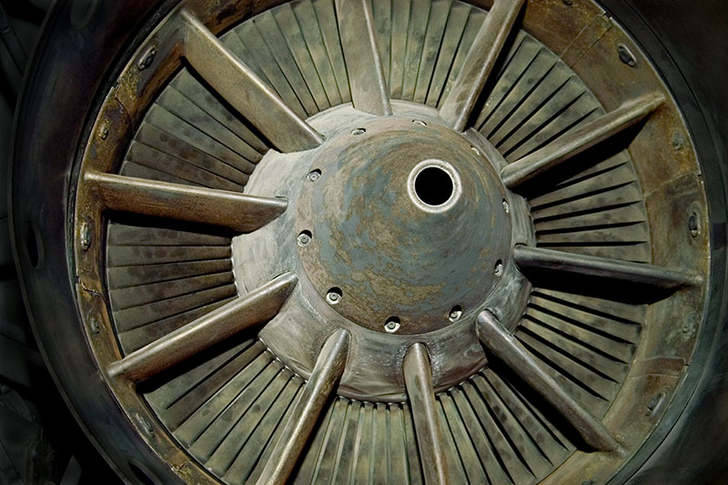 Close-up view of a weathered metal turbine or fan with radial blades arranged in a circular pattern around a central hub. The surface shows signs of age and oxidation with rust-colored patina, while the geometric arrangement of the flat, rectangular blades creates a striking symmetrical composition against the dark background.