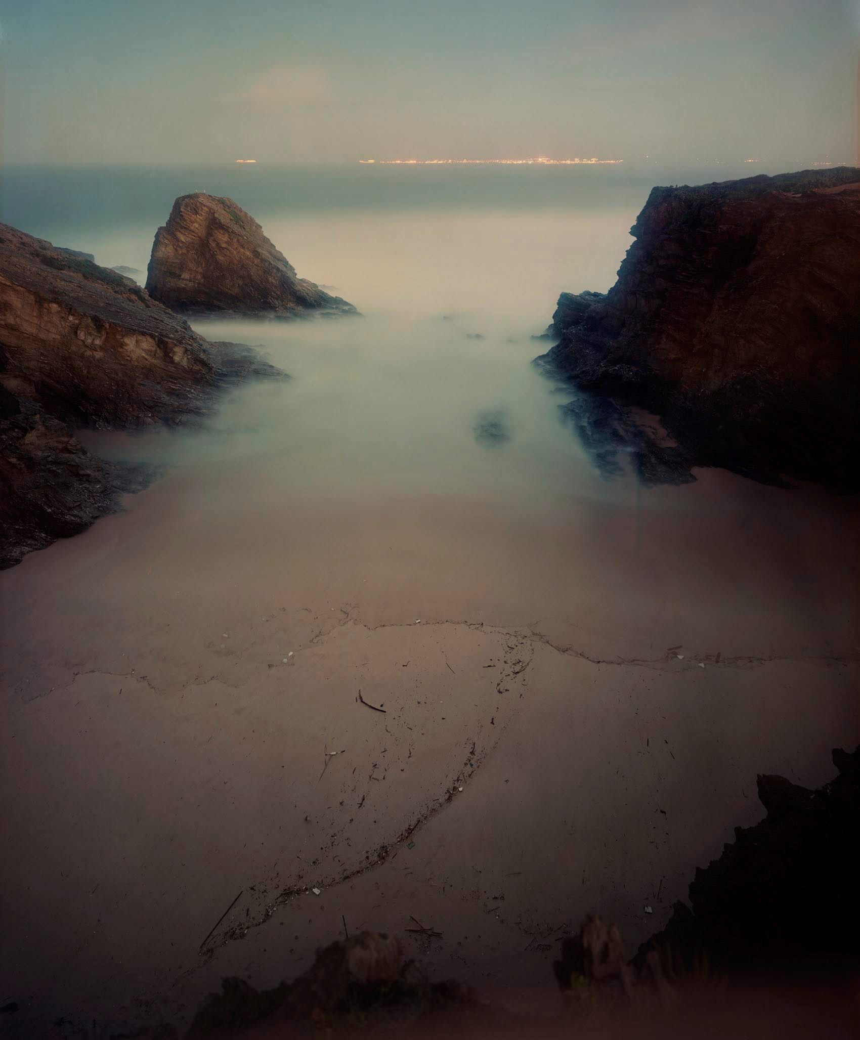 Misty coastal landscape with a sandy beach in the foreground marked by a spiral pattern, flanked by dramatic dark rocky outcrops on either side. The seawater blurs into soft, ethereal tones in muted blues and grays, with distant lights visible along the horizon beneath a hazy sky.