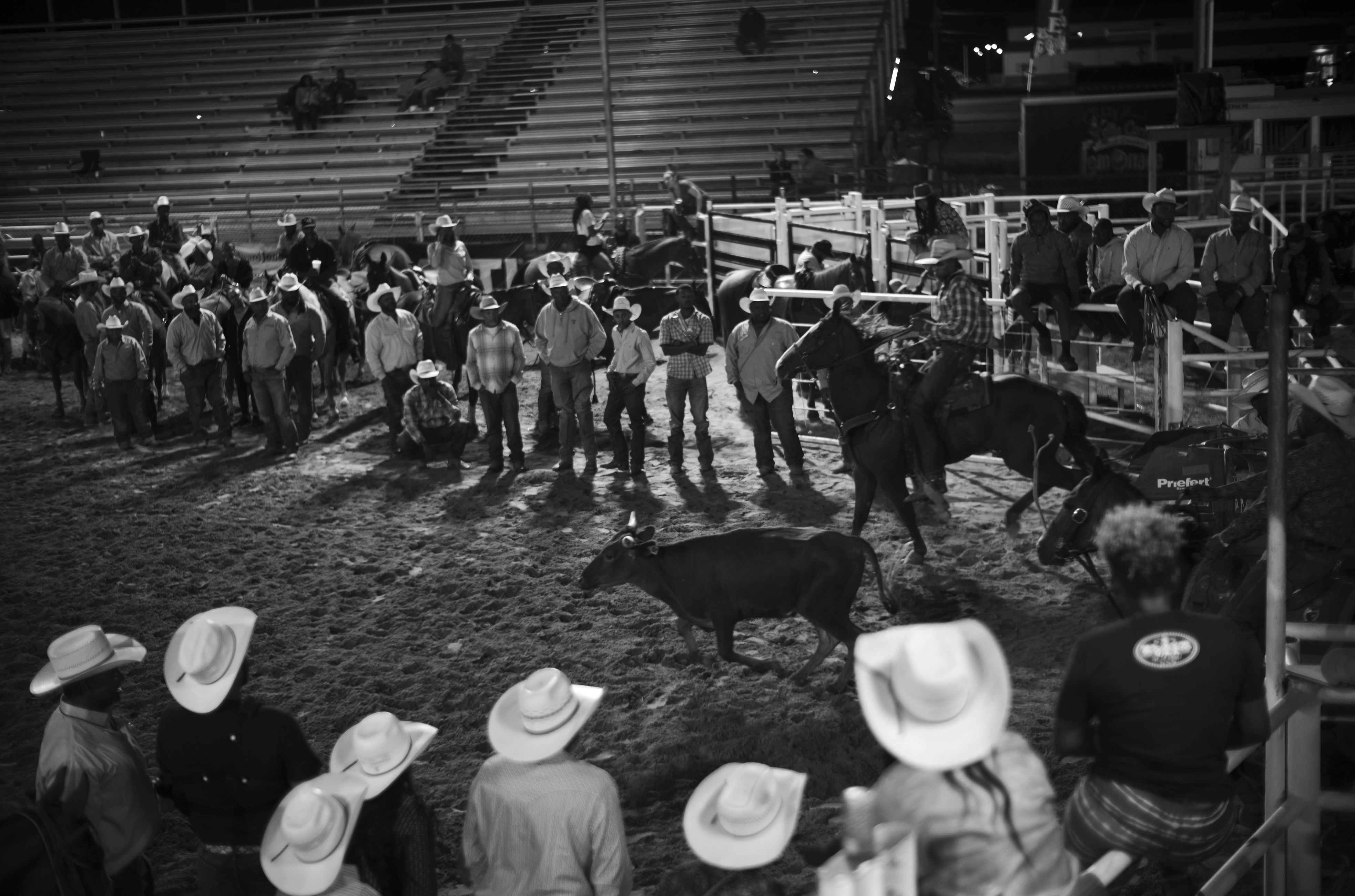 Black and white photograph of a rodeo arena scene with cowboys wearing wide-brimmed hats gathered in the foreground and background, with wooden bleachers and fencing visible. A bull stands in the center of the sandy arena surrounded by the spectators and participants.