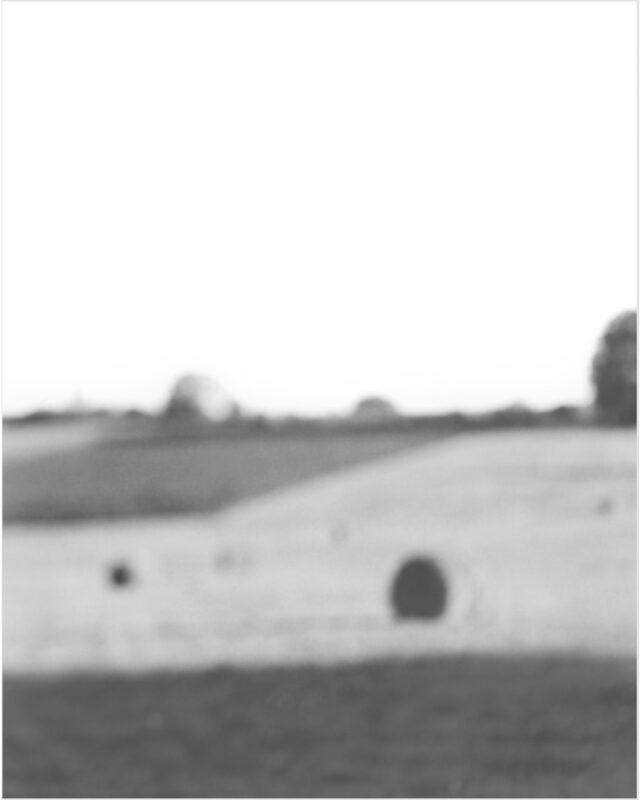 Black and white photograph of a blurred landscape with a flat field in the foreground and trees along the horizon beneath a pale sky. Two dark circular forms appear in the middle ground against the lighter terrain.
