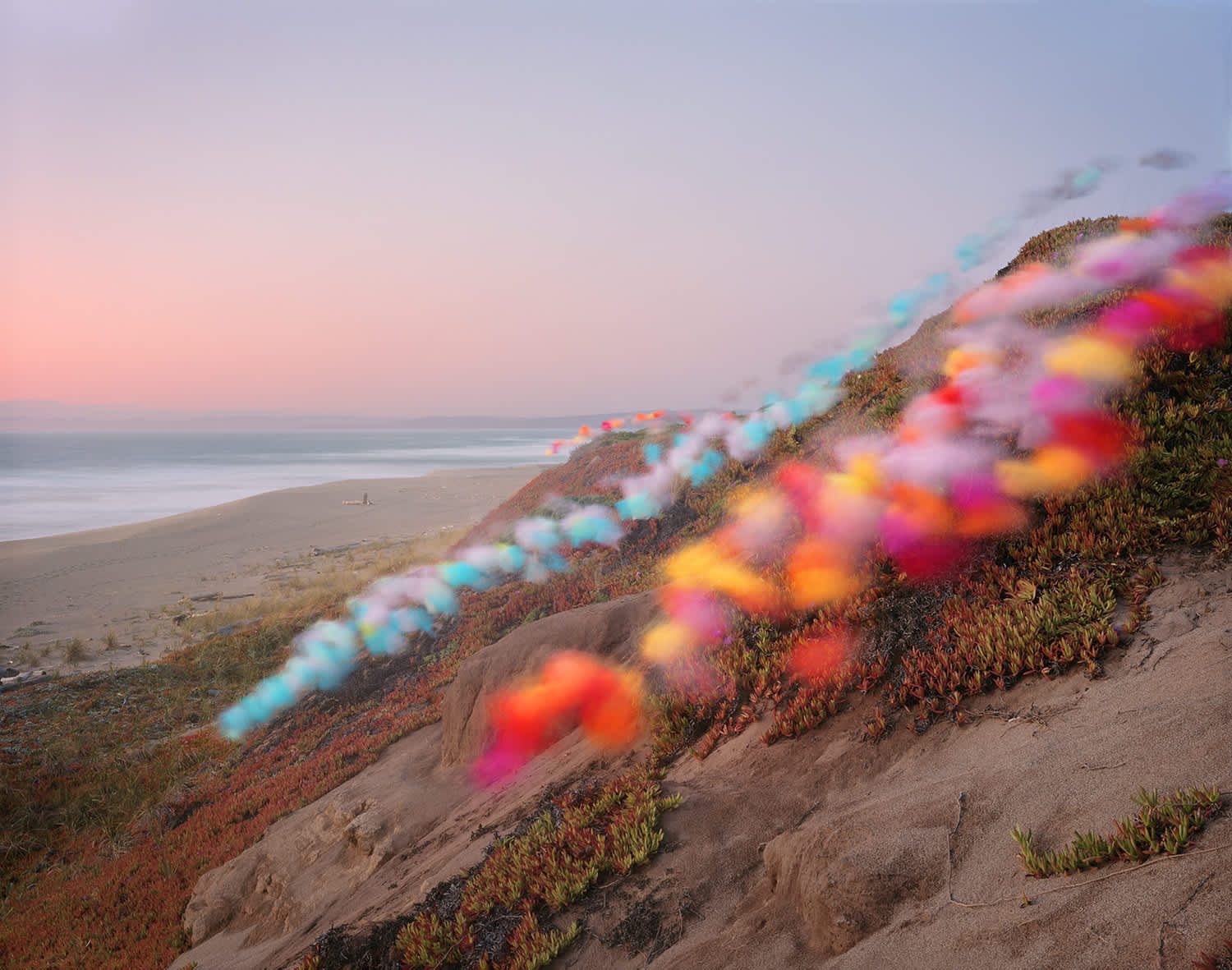 Coastal landscape at dusk featuring a sandy cliff overlooking a beach and ocean, with a diagonal line of vibrant, blurred colored powder or pigment—in shades of cyan, magenta, yellow, and orange—cascading down the hillside against the muted tones of the terrain and twilight sky.
