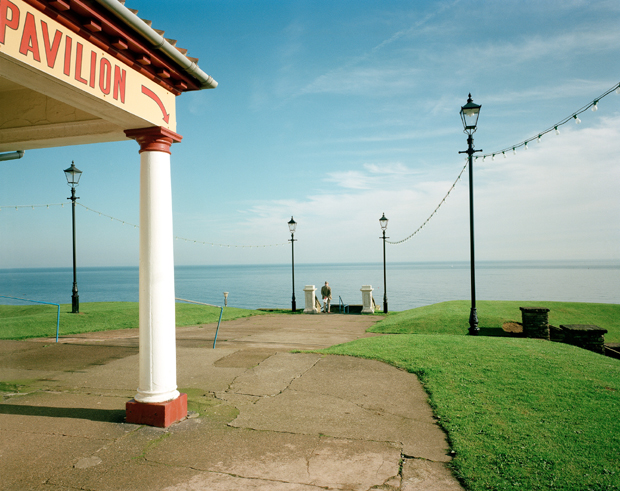 A seaside pavilion with a white column and red trim stands in the foreground, its signage partially visible. Beyond it, manicured green lawns slope toward a calm sea under a pale blue sky, with vintage street lamps and string lights creating a nostalgic atmosphere along the coastal promenade.