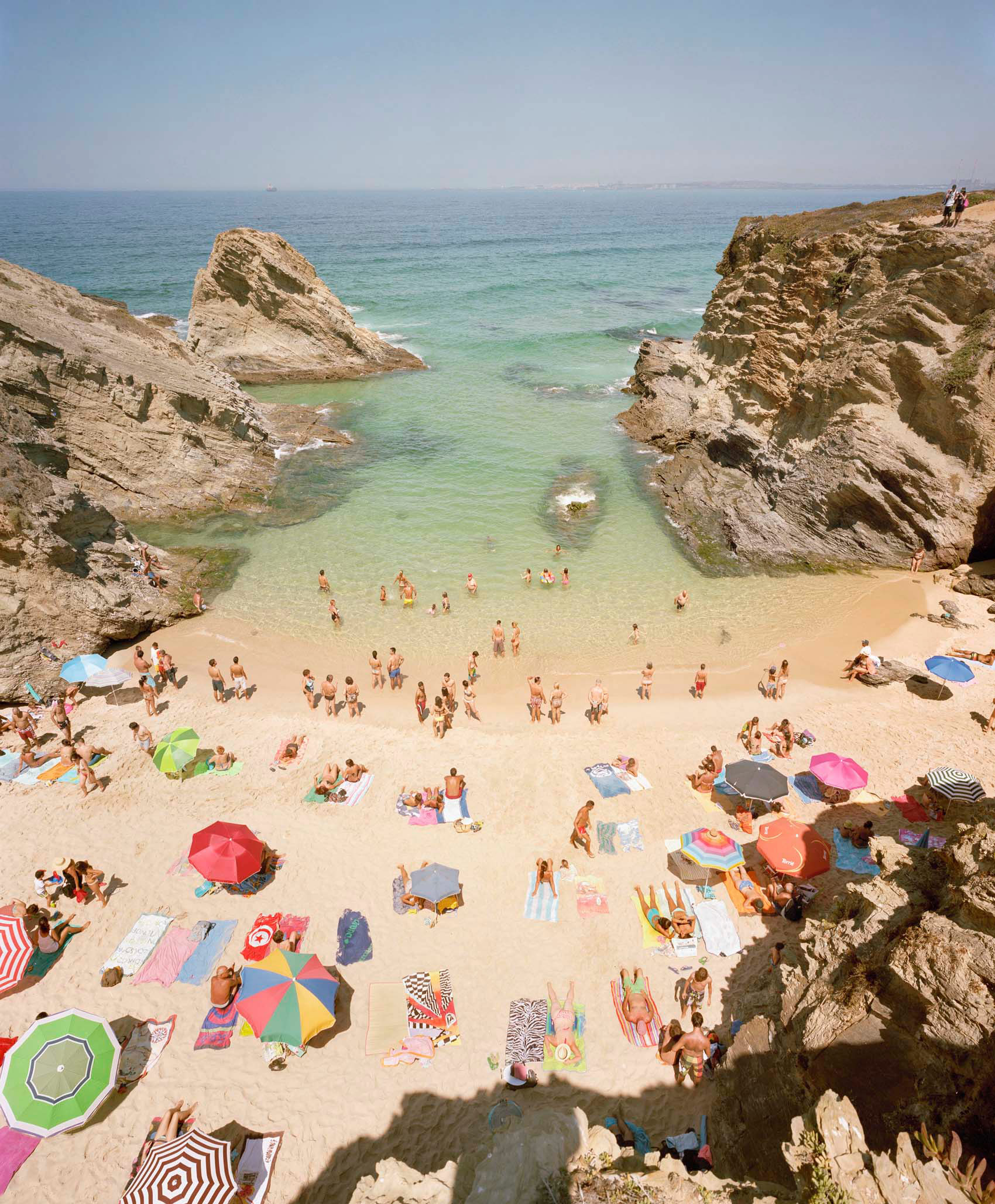 Aerial view of a crowded beach nestled between dramatic limestone cliffs, with colorful umbrellas and beach towels dotting the golden sand, turquoise water, and numerous swimmers and sunbathers scattered across the scene.