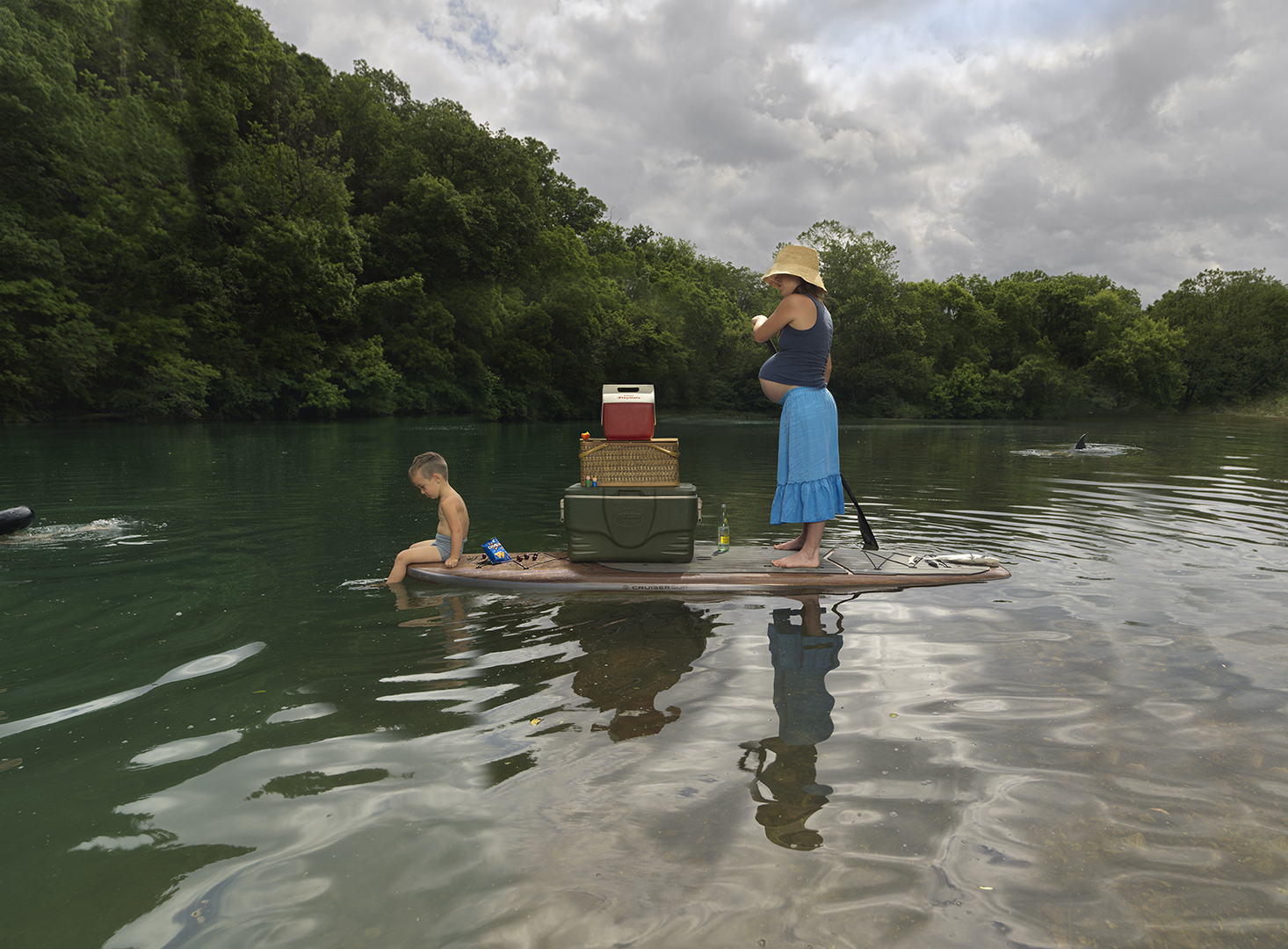 A woman in a straw hat and blue skirt stands on a paddleboard in shallow water, holding a paddle while looking toward a young child wading nearby. A cooler and picnic basket sit between them on the board, with forested riverbanks and cloudy sky reflected in the calm water's surface.