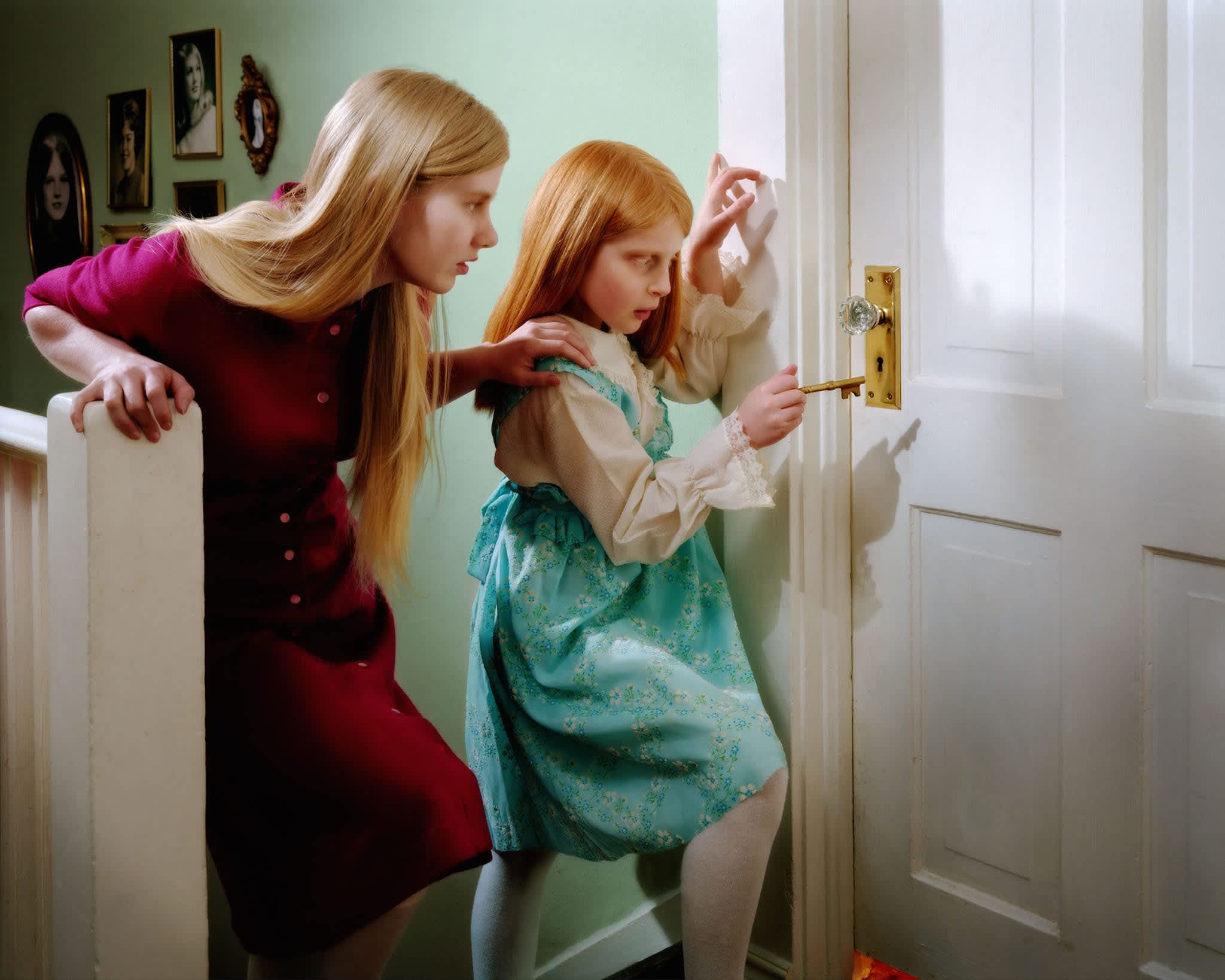 A woman in a burgundy dress leans over a young girl in a turquoise floral dress as the child reaches toward a brass doorknob on a white door. Set against a soft sage green wall, the composition captures a moment of curiosity and gentle guidance, rendered in rich, saturated color with hyperrealistic detail.