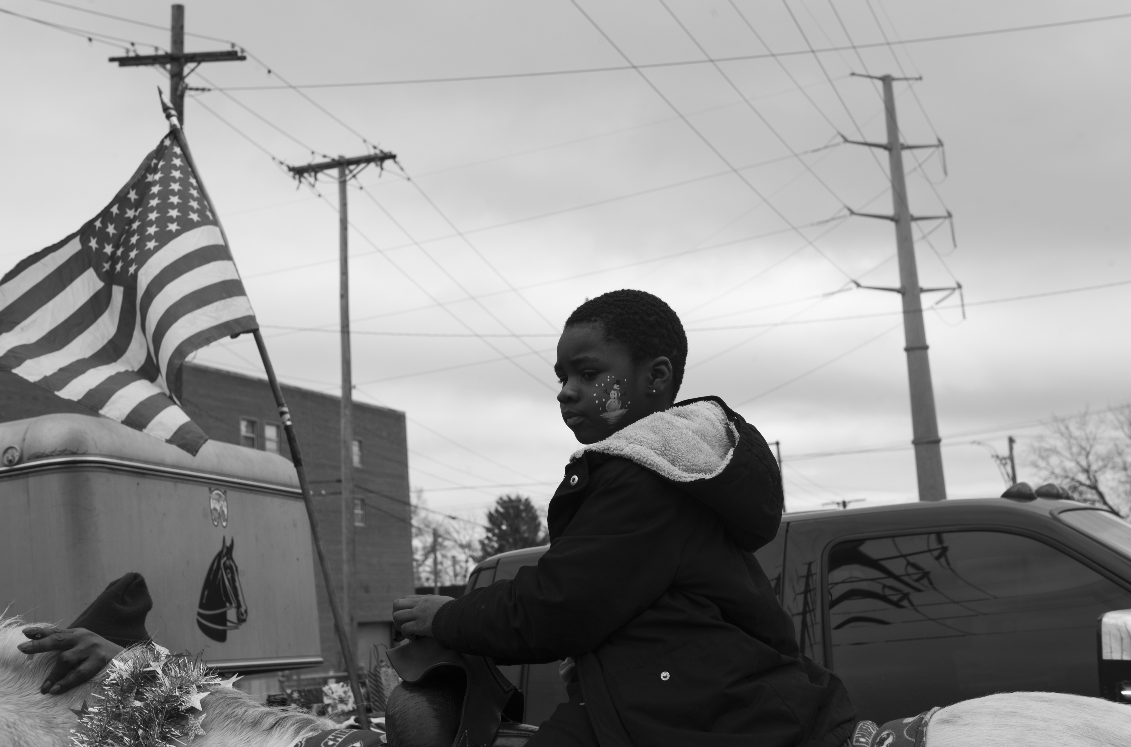 Black and white photograph of a young child sitting in a vehicle, looking to the side. An American flag waves from the vehicle's antenna in the background, with utility poles and overcast sky visible behind. The composition captures an intimate moment within an industrial American landscape.
