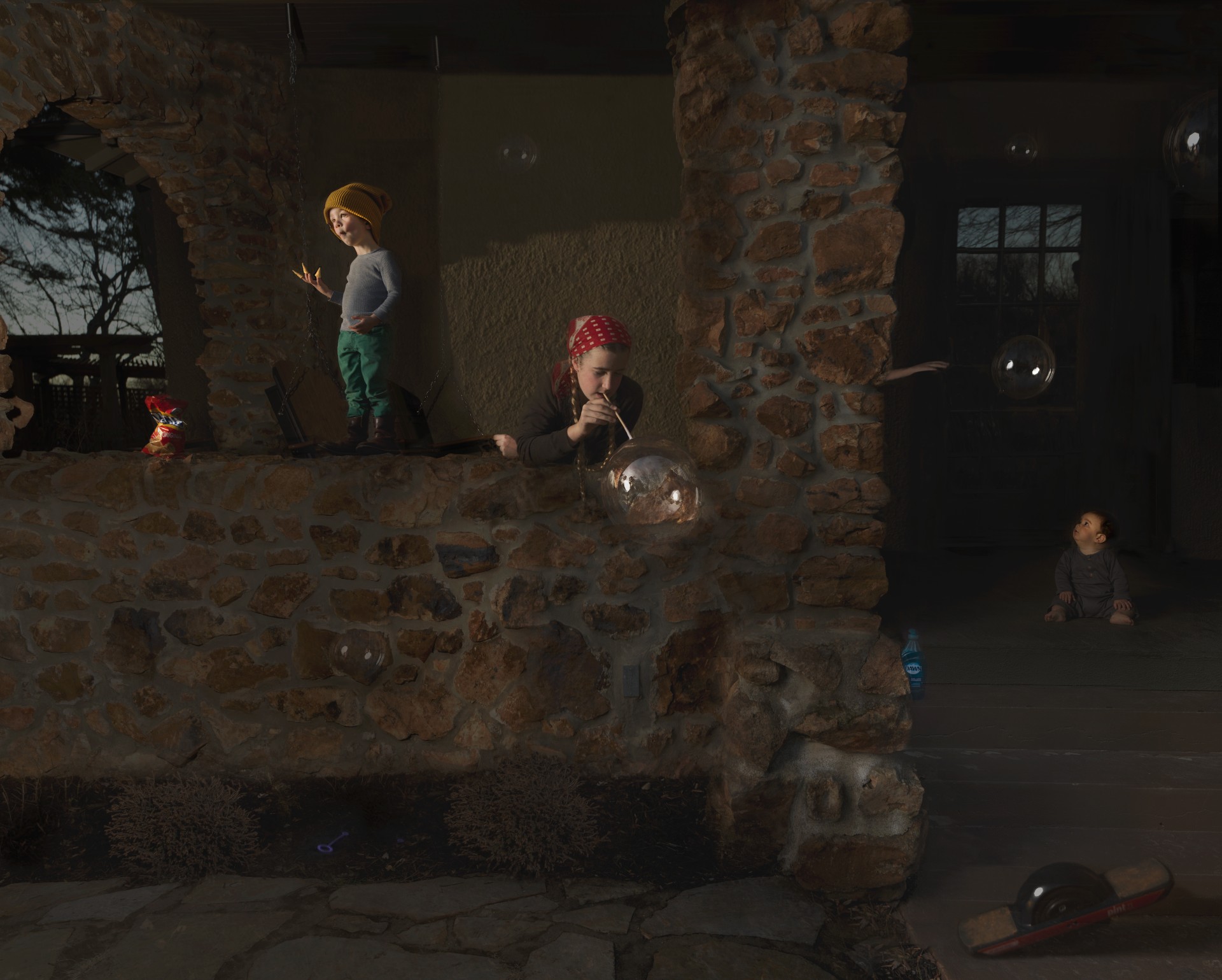 Three children play in a stone-walled interior space illuminated by dramatic natural light. One child in a yellow hat stands to the left, another wearing a red bandana blows soap bubbles in the center, and a third sits near a doorway on the right. The warm, golden light casts strong shadows across the weathered brick and stone surfaces, creating a contemplative atmosphere in this domestic scene.