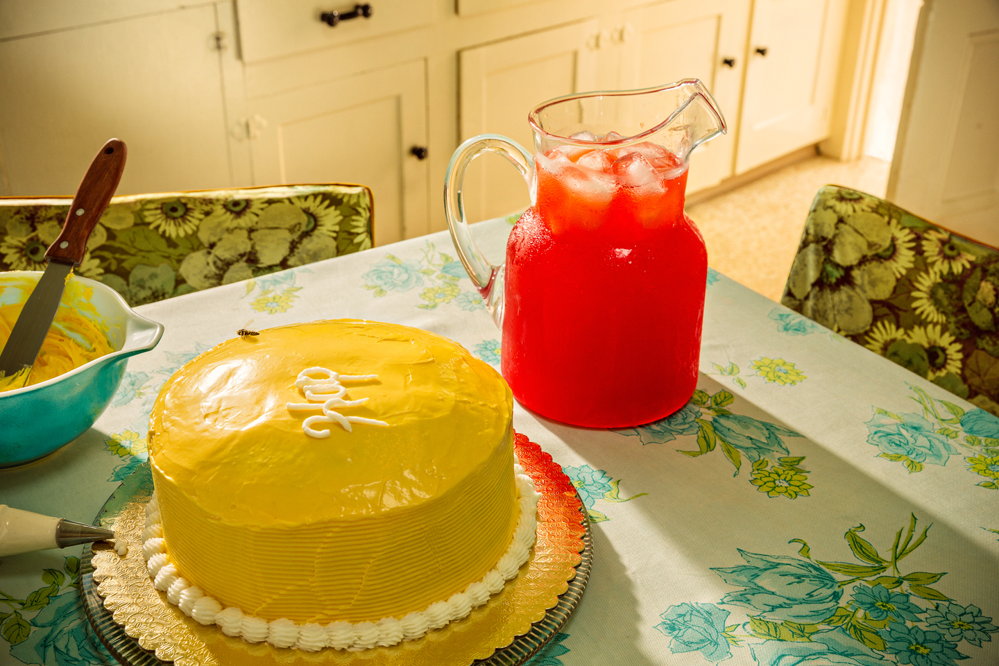 A bright yellow cake with white piped frosting border sits on a gold cake board alongside a glass pitcher filled with red beverage and ice cubes. The table is set with a floral patterned tablecloth in yellows and greens, with green-upholstered chairs visible in the background and cream-colored kitchen cabinets beyond.