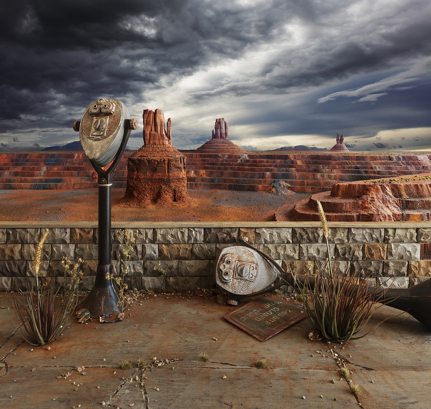 Two weathered coin-operated viewing telescopes stand on a stone wall overlooking a vast desert landscape with distinctive red rock formations and buttes. Dramatic storm clouds gather overhead while desert vegetation and a worn plaque rest on the ground below, creating a scene that blends authentic monument viewpoint imagery with surreal, constructed elements.