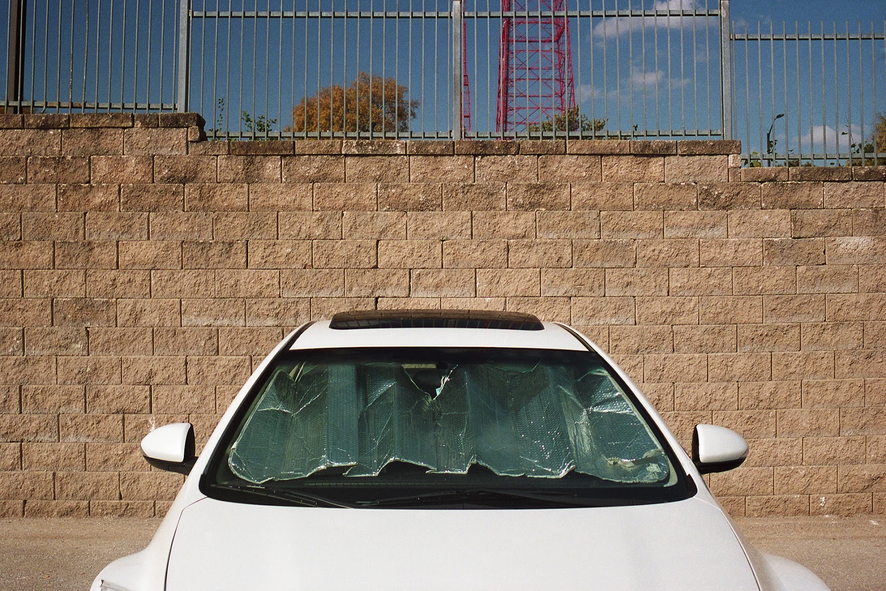 White sedan parked against a tan brick wall with a green sunshade covering its windshield, industrial metal fencing and red transmission tower visible in the background under a blue sky.