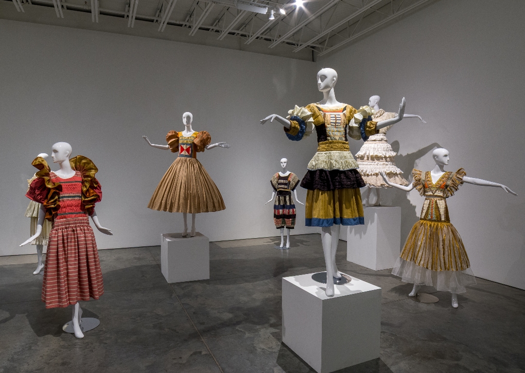 Seven mannequins display elaborate fiber garments in a gallery space, featuring hand-pleated skirts, embroidered details, and voluminous shoulders in rich jewel tones and gold metallics. The sculptural garments, positioned on white pedestals against gray walls, blend historical costume references with contemporary textile techniques including patchwork and hand-stitching.