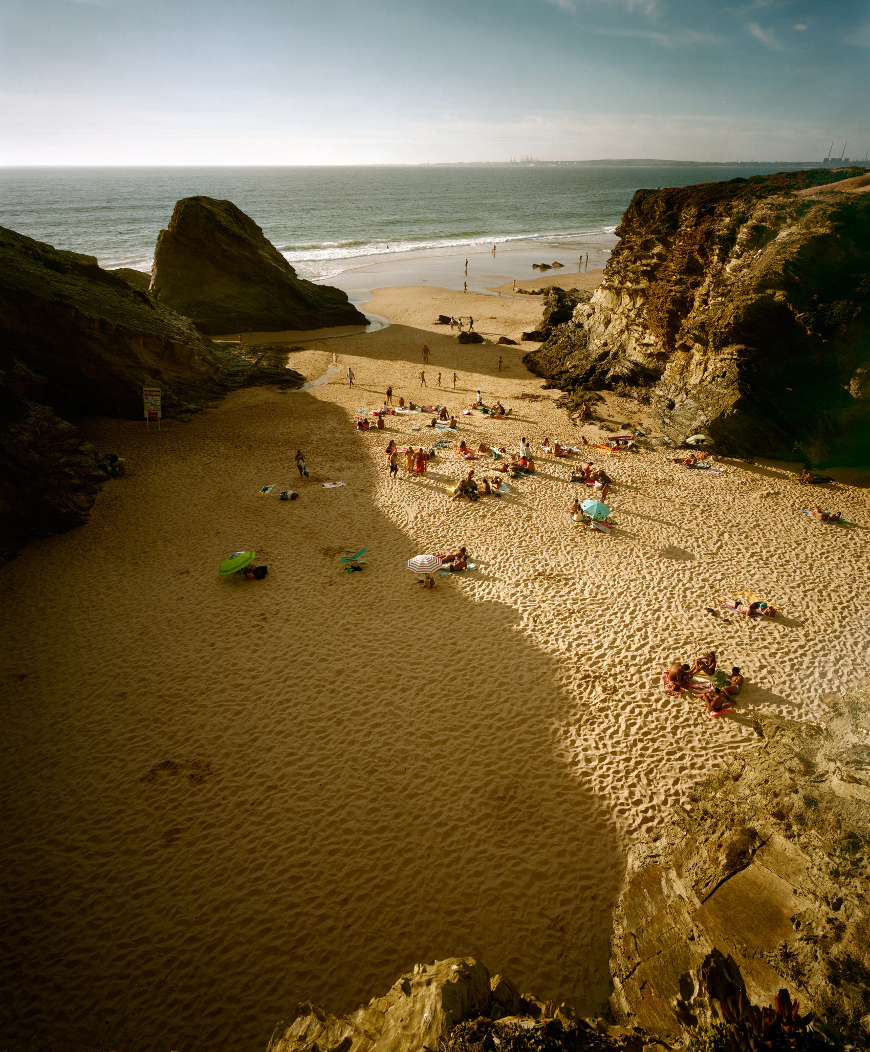 Aerial view of a sheltered beach cove framed by dramatic rocky cliffs, with scattered beachgoers on pale sand and shallow water. Sailboats visible on the distant horizon beneath a hazy sky.