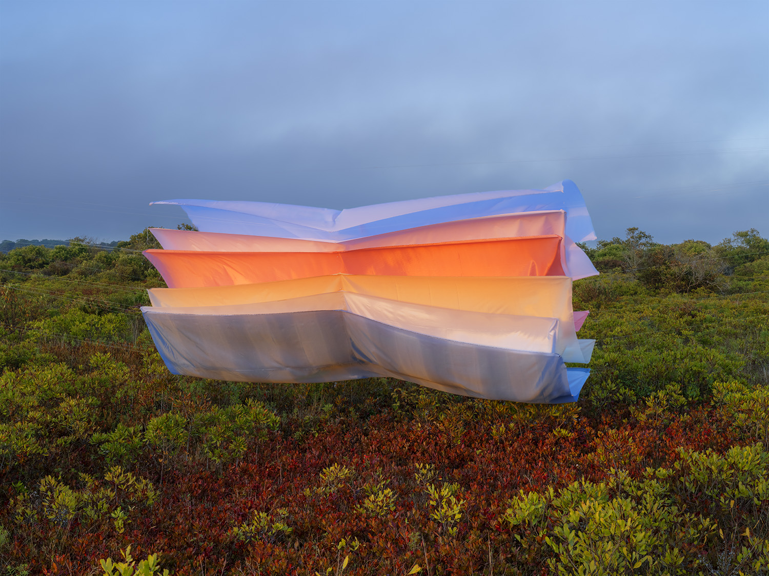 Layered sheets of translucent colored acetate in blue, orange, yellow, and white are suspended above low scrubland vegetation, their luminous surfaces catching light against an overcast sky.
