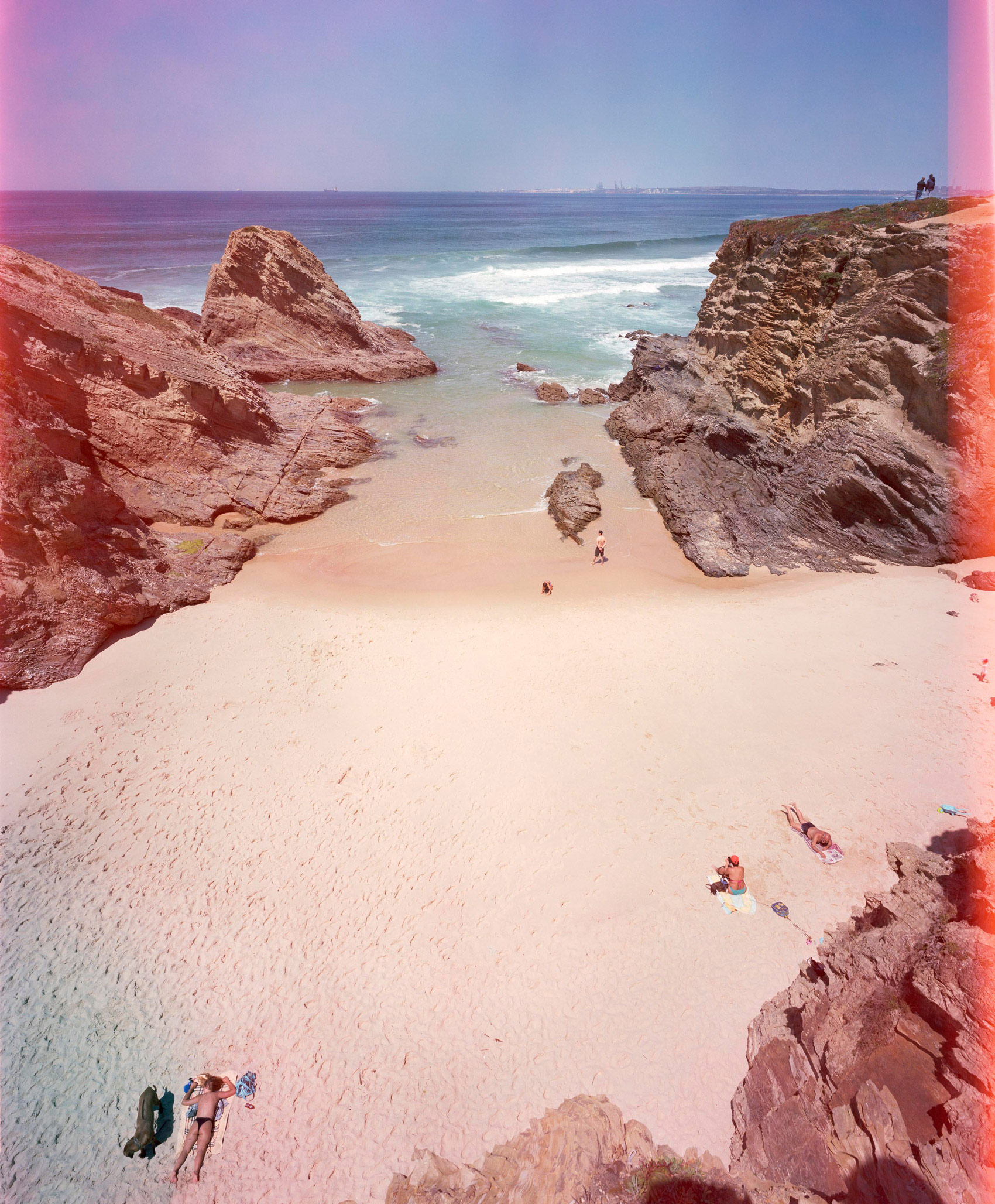 A surreal beach cove framed by towering red rock formations, with pale pink sand and turquoise waters. Small figures of beachgoers dot the pristine shore, while the sky and landscape are saturated in warm magenta and cool blue tones, creating a dreamlike, color-shifted atmospheric effect.