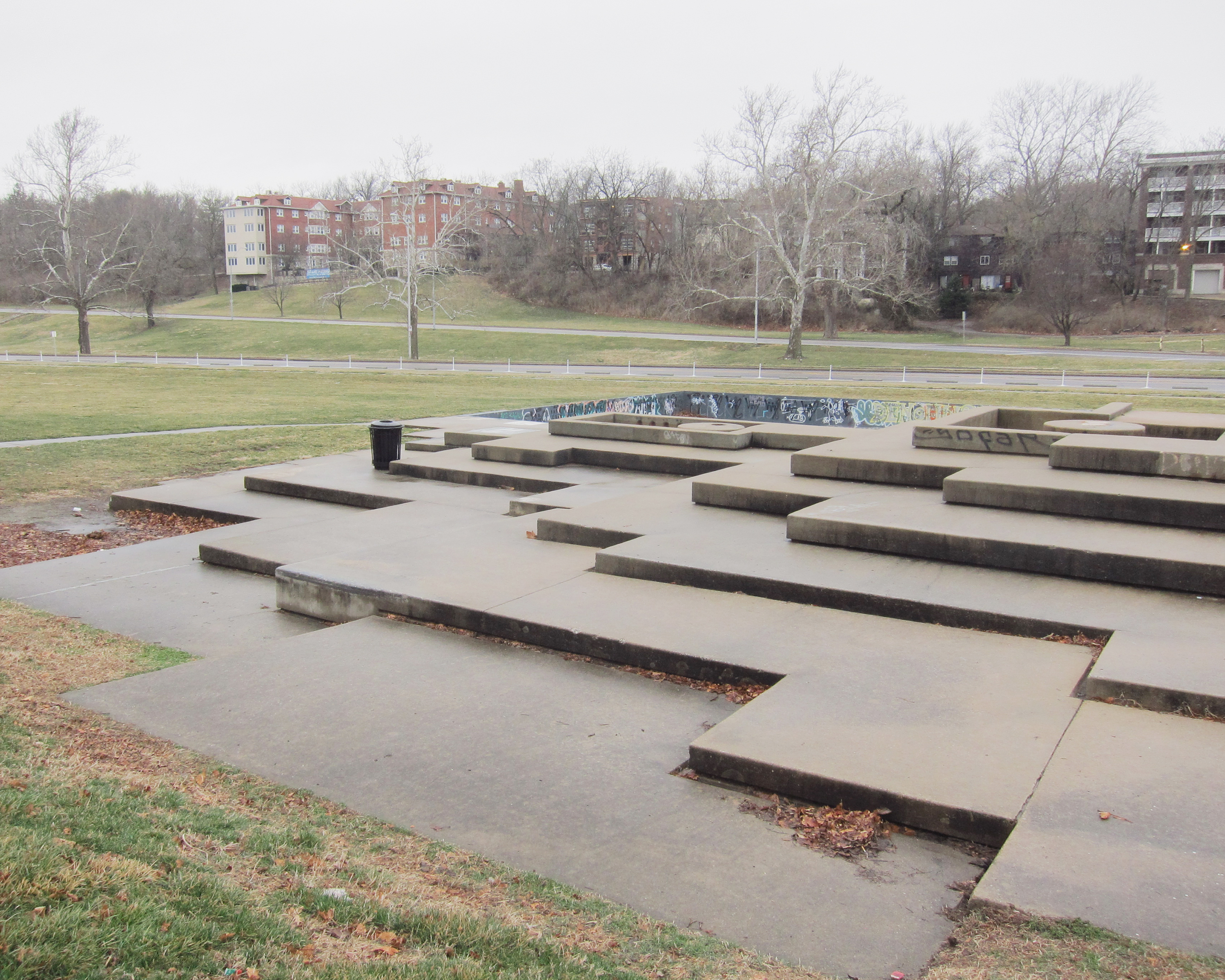 Concrete platforms and stepped forms of varying heights arranged across a paved plaza in an urban park, with bare trees and brick apartment buildings visible in the background under an overcast sky.