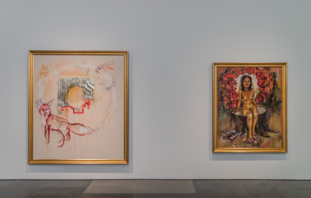 Two framed paintings hang on a gallery wall separated by white space. On the left, a larger painting features abstracted figures in red and orange tones with decorative line work on a cream background. On the right, a smaller painting depicts a seated figure in gold and yellow clothing centered within a vibrant red floral wreath against a darker background. Both works are mounted in gold frames and appear designed as backdrops for viewer interaction.